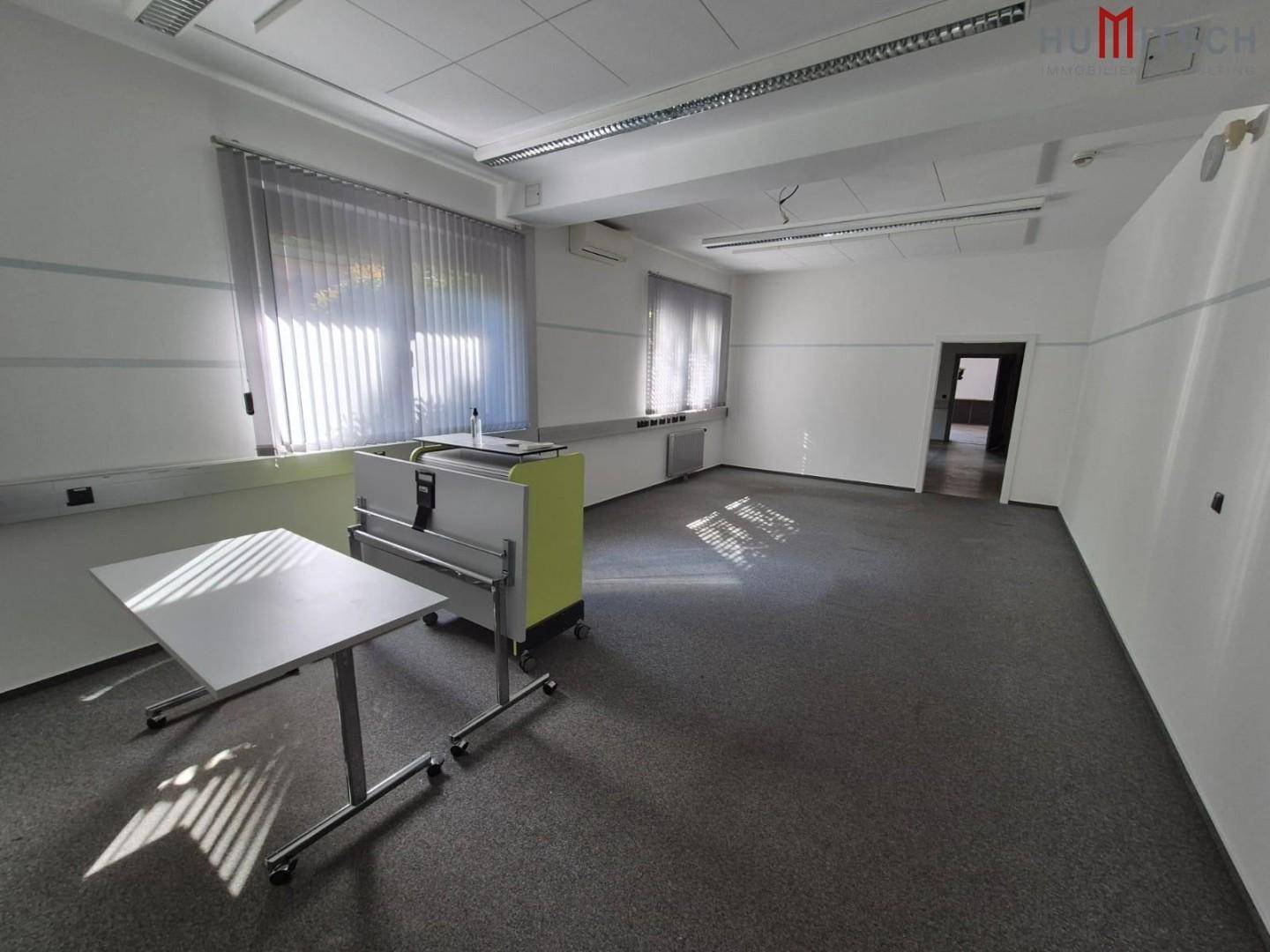 Helles Büro mit Teppichboden, mobilen Tischen und großen Fenstern.