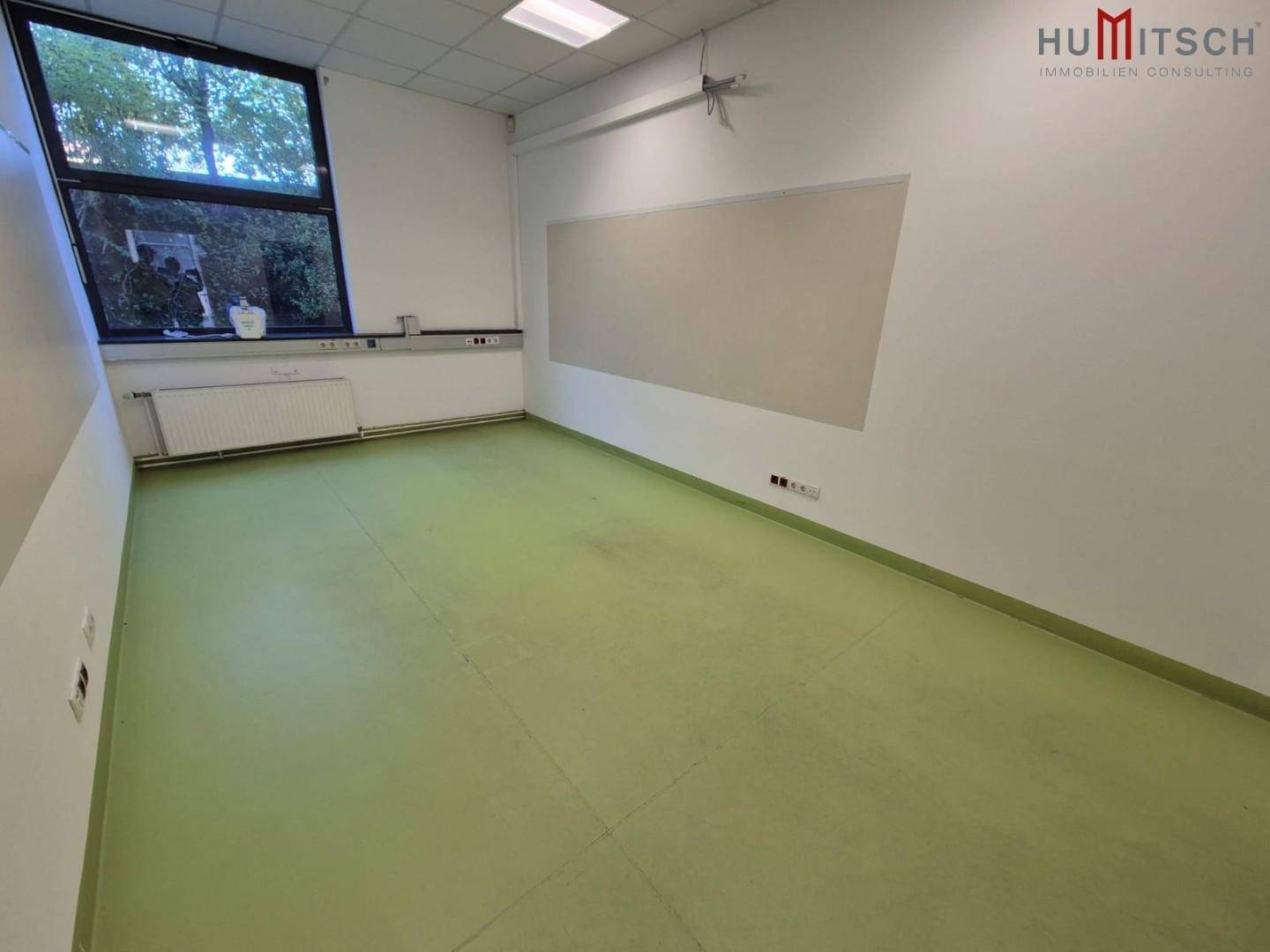 Helles Büro mit grünem Linoleumboden, großen Fenstern und einer Tafel.