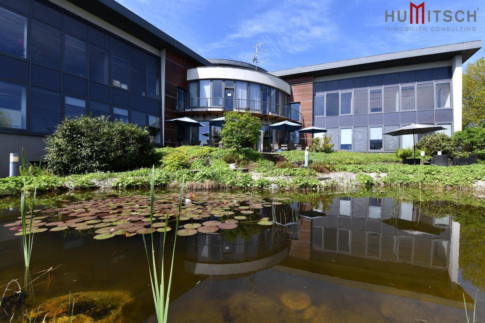 Modernes Bürogebäude mit großen Glasfassaden und einem Teich im Vordergrund unter blauem Himmel.
