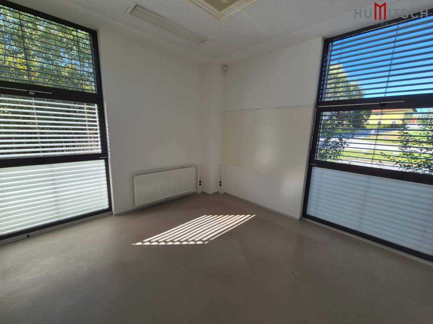 Büroraum mit grauem Boden, großen Fenstern mit Jalousien und viel Tageslicht.