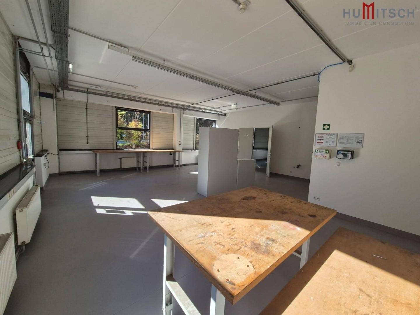 Großer Arbeitsraum mit mehreren Tischen, sichtbaren Leitungen und Fenstern mit Blick ins Grüne.