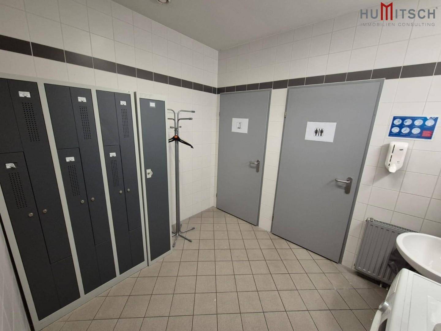 Umkleideraum mit Fliesenboden, Spinden und zwei Türen zu Sanitärbereichen.