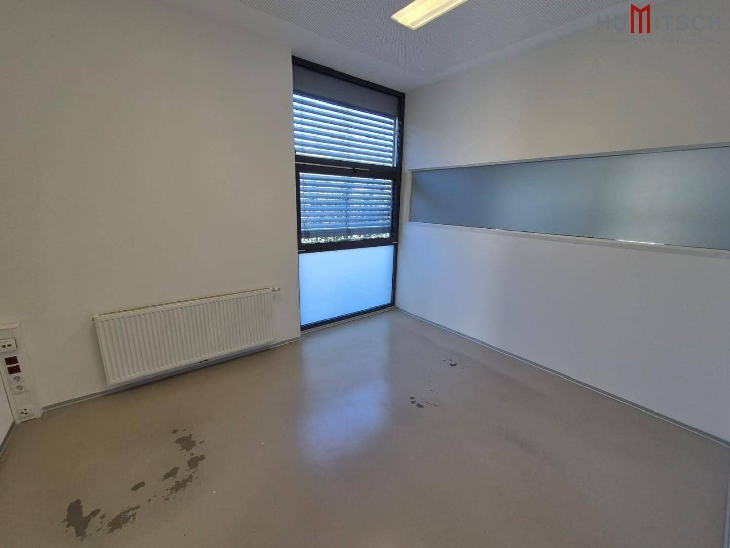 Büroraum mit hellem Boden, großem Fenster mit Jalousien und Heizkörper.