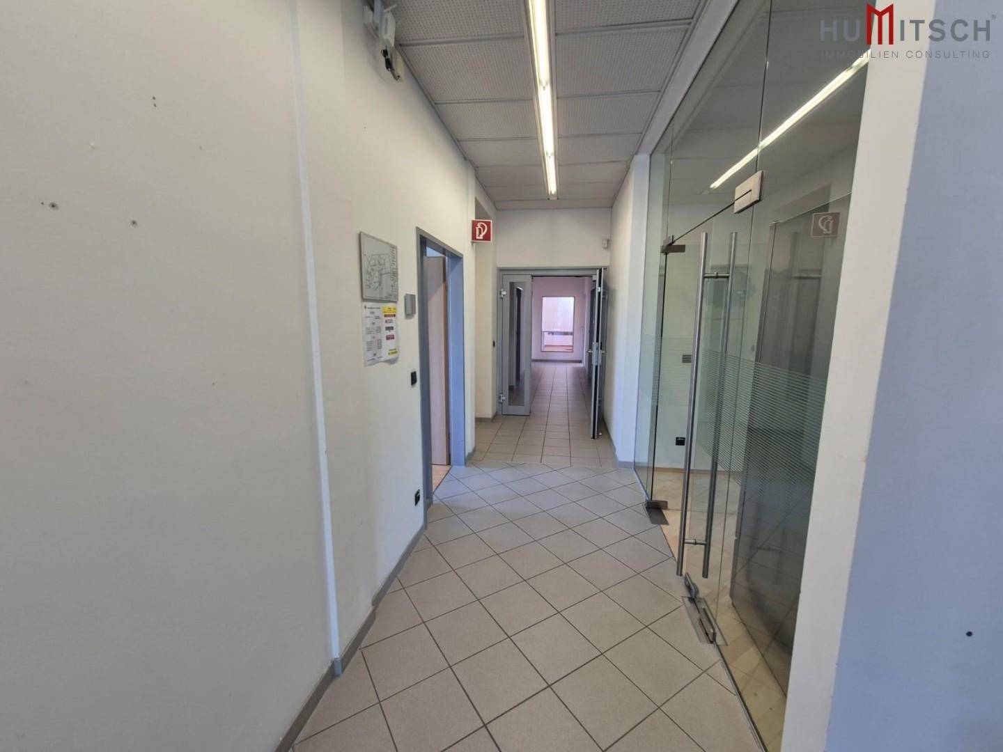 Langer Flur mit Fliesenboden, weißen Wänden und Glastüren zu Büroräumen.