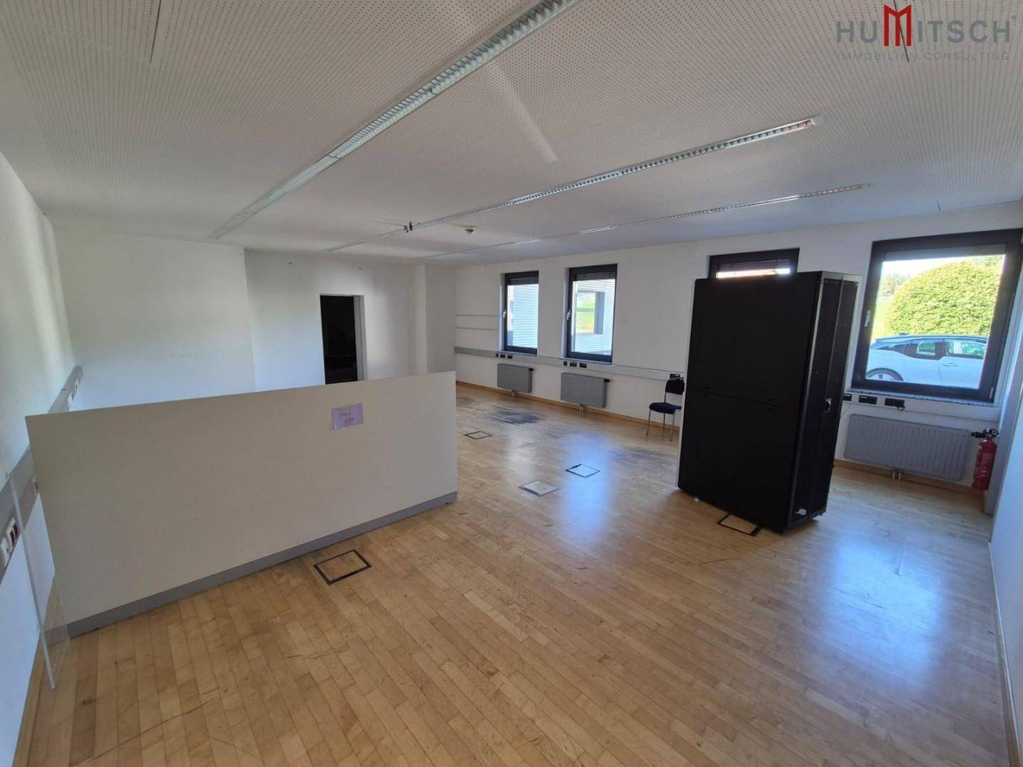 Heller Büroraum mit Holzparkett, Trennwand und Fenstern mit Blick ins Grüne.