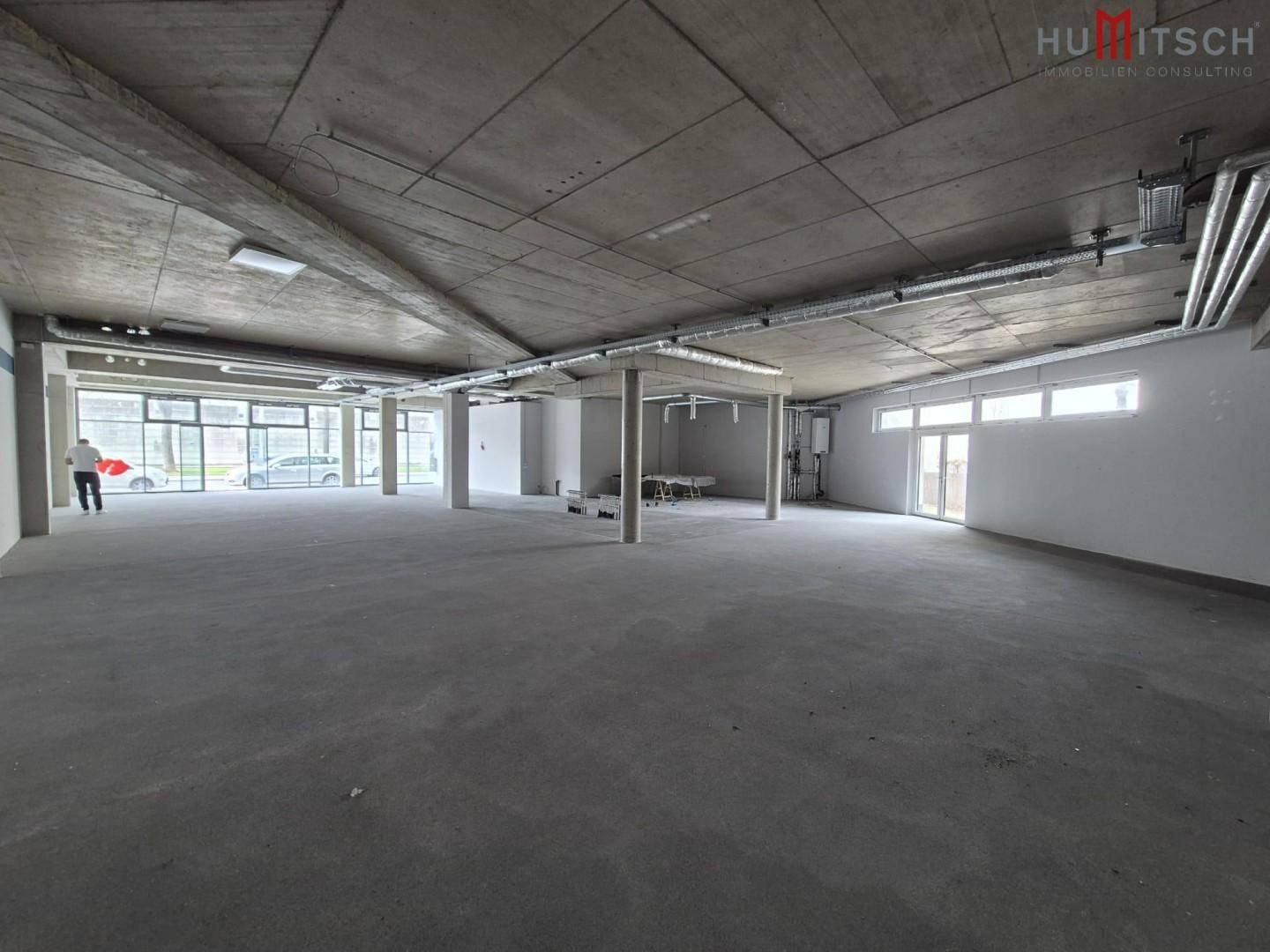 Weitläufiger Gewerberaum mit Sichtbeton und viel Tageslicht durch große Fensterfronten.