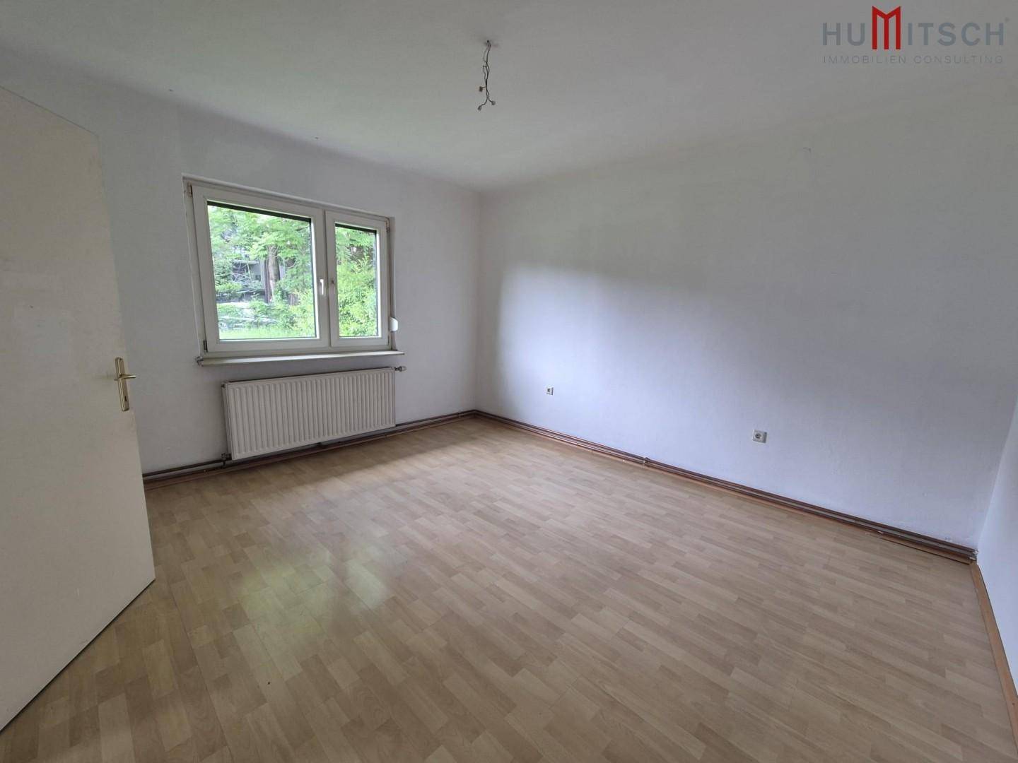 Leeres Schlafzimmer mit hellem Laminatboden, weiß gestrichenen Wänden und einem Fenster mit Blick ins Grüne.