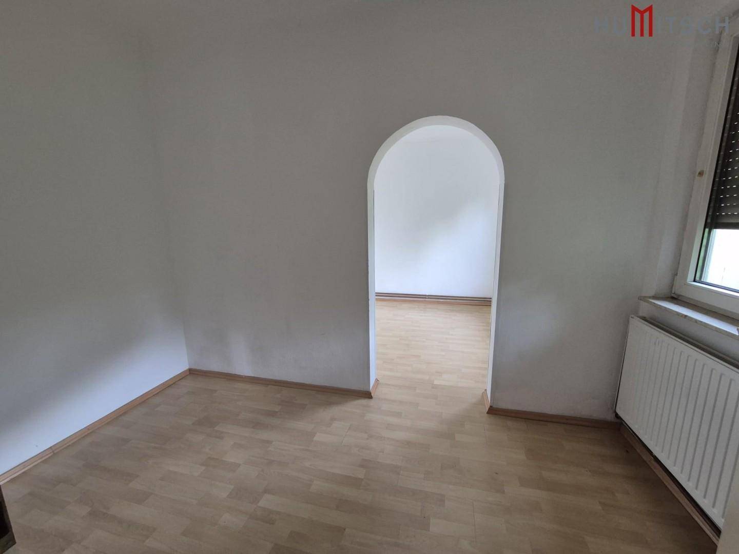 Heller Raum mit Laminatboden und einem Rundbogen, der zu einem weiteren Zimmer führt.