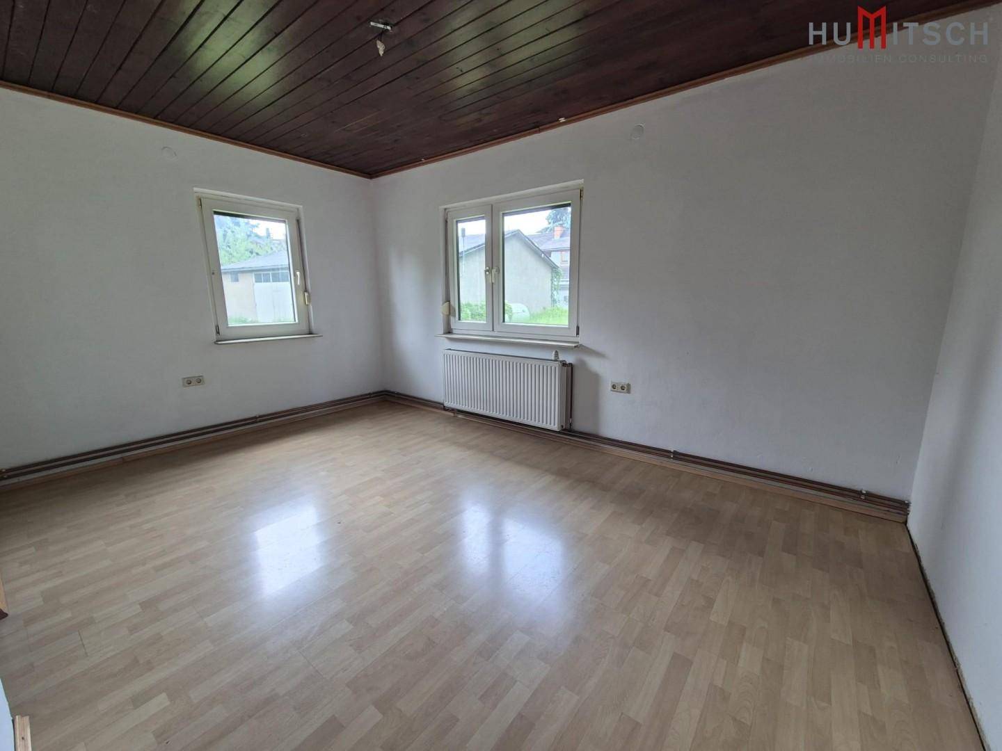 Unmöbliertes Zimmer mit Holzbalkendecke, Laminatboden und zwei Fenstern, die Tageslicht hereinlassen.