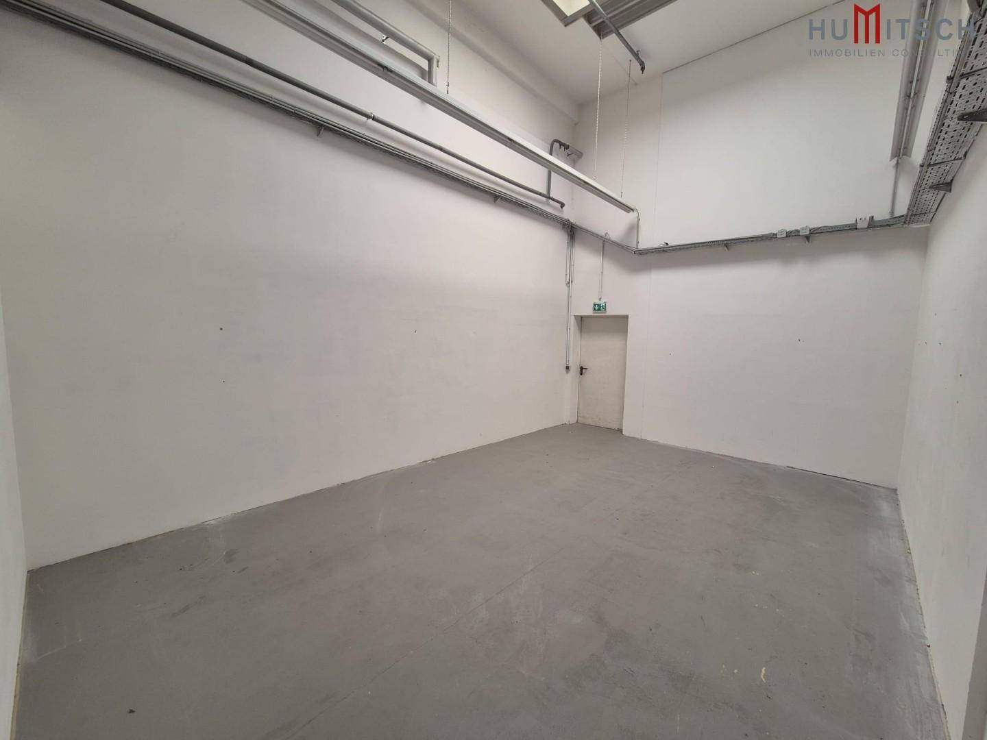 Einfacher Lagerraum mit Betonboden und hoher Decke, ausgestattet mit einer Notausgangstür.