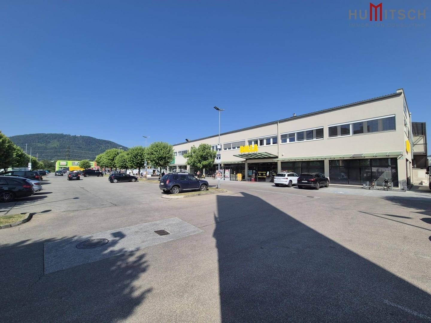 Eingangsbereich eines modernen Gewerbegebäudes mit Parkplätzen unter strahlend blauem Himmel.