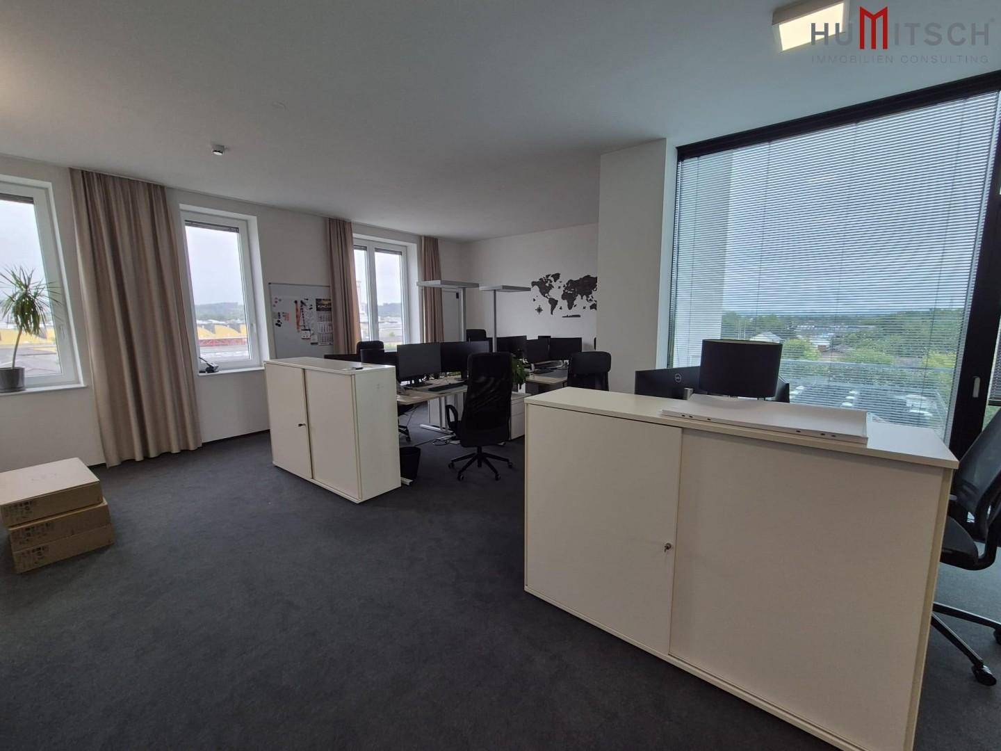 Großraumbüro mit mehreren Arbeitsplätzen, weißen Schränken und großen Fenstern.