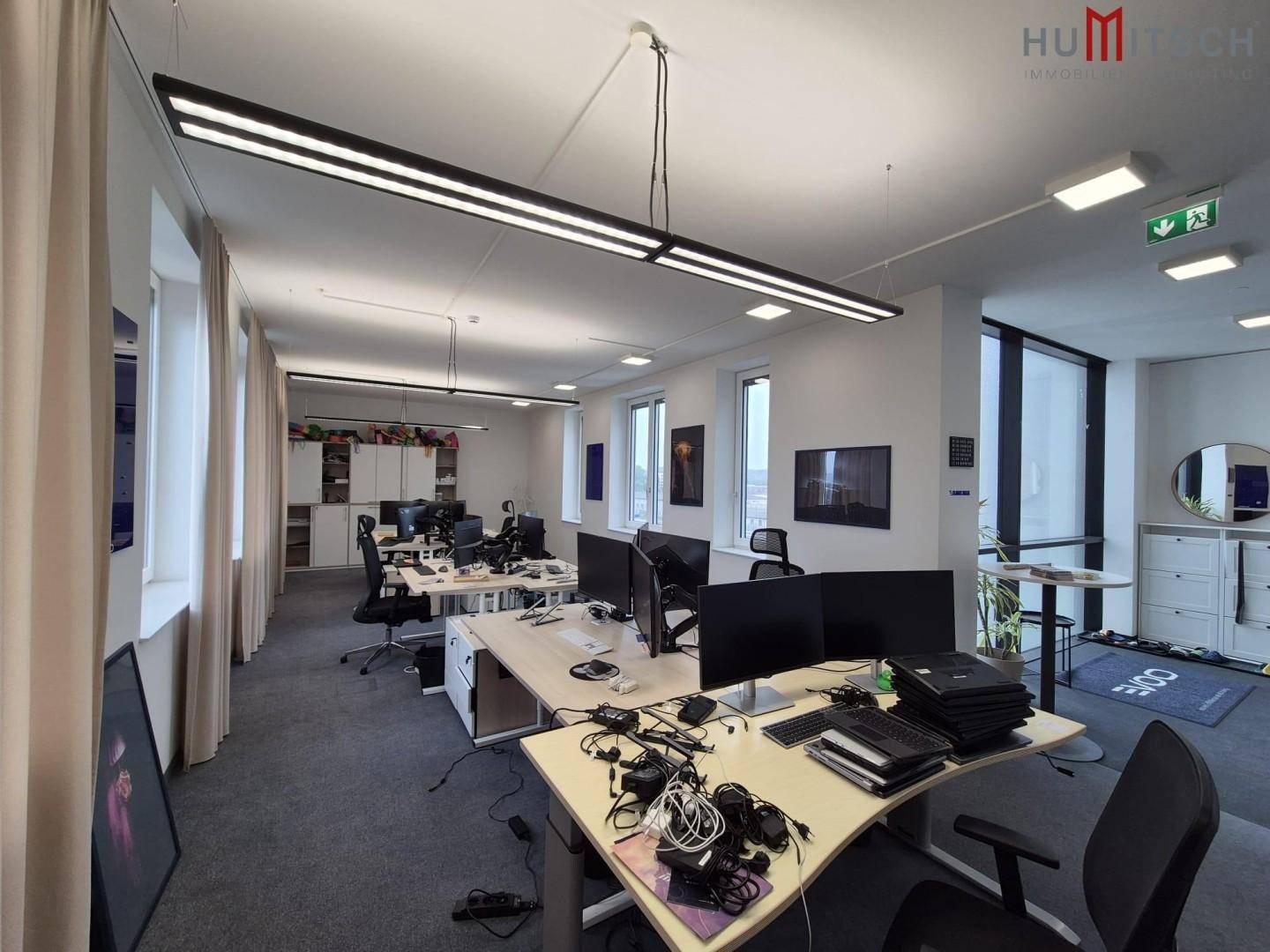 Offenes Büro mit vielen Arbeitsplätzen, Teppichboden und modernen Hängelampen.