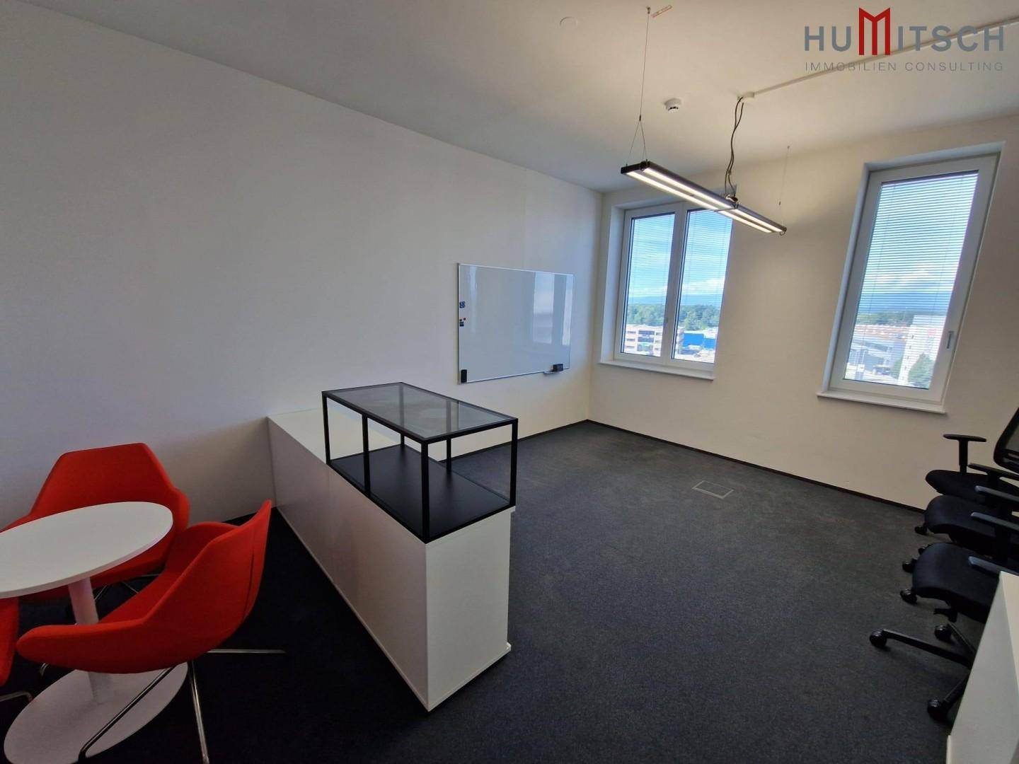 Büroraum mit Teppichboden, großen Fenstern und einer kleinen Sitzecke mit roten Stühlen.