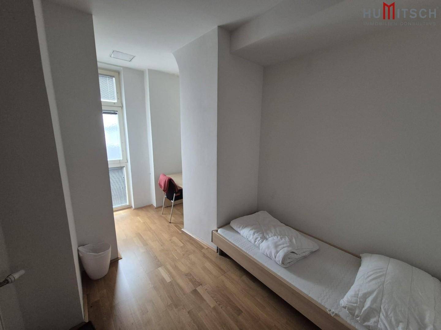 Schlichtes Schlafzimmer mit Einzelbett und Blick auf einen Arbeitsbereich durch einen Durchgang.