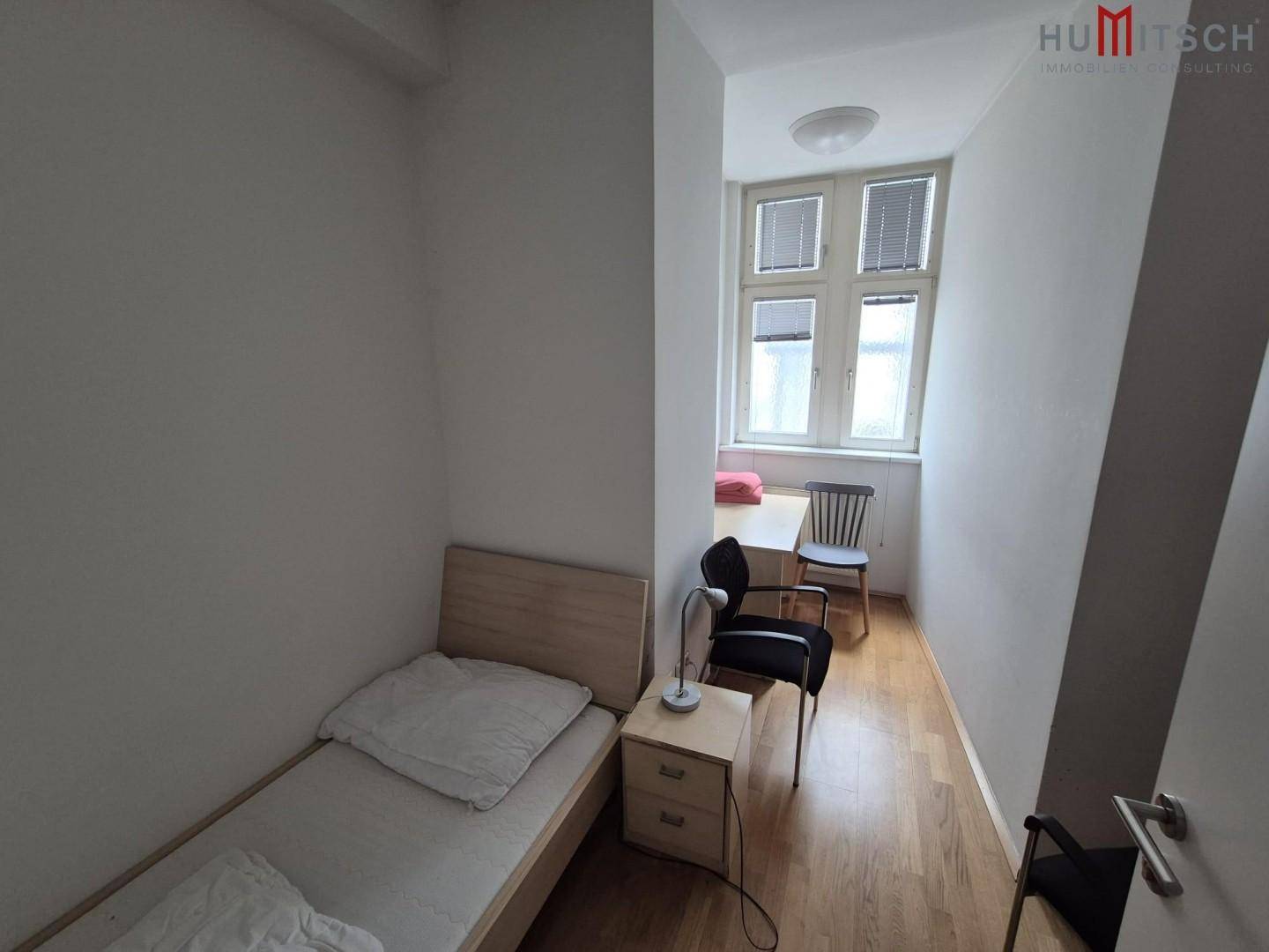 Schlichtes Schlafzimmer mit Einzelbett und Schreibtisch, hell durch ein großes Fenster.