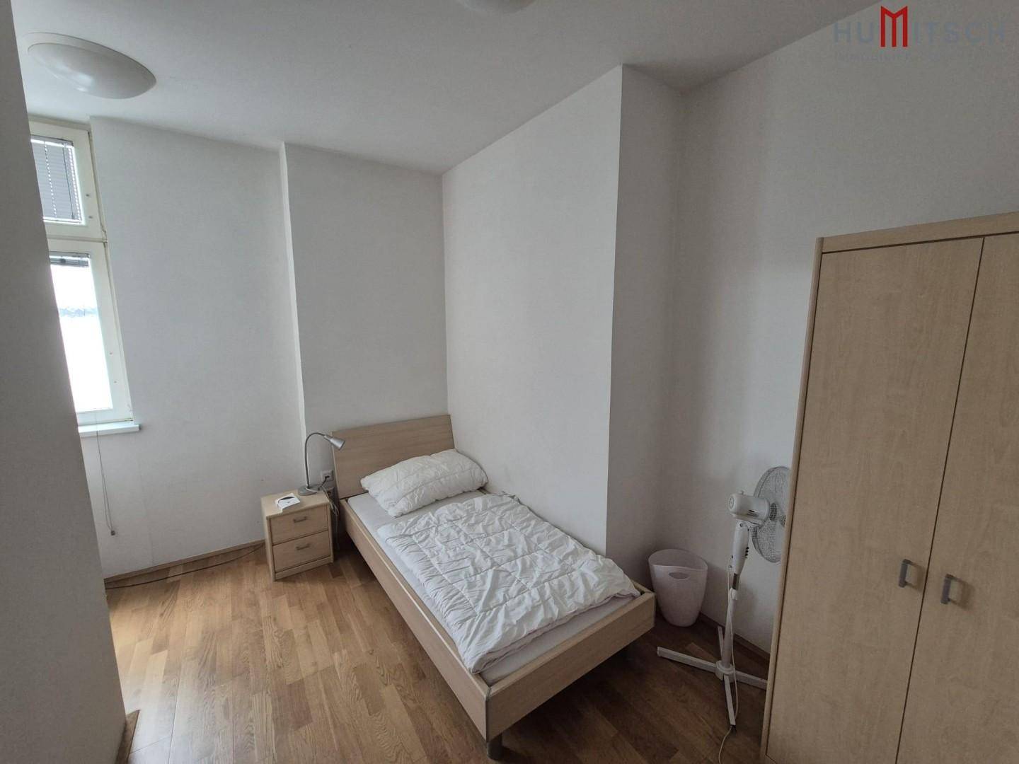 Helles Schlafzimmer mit Einzelbett, Nachttisch und Kleiderschrank, ausgestattet mit einem Ventilator.