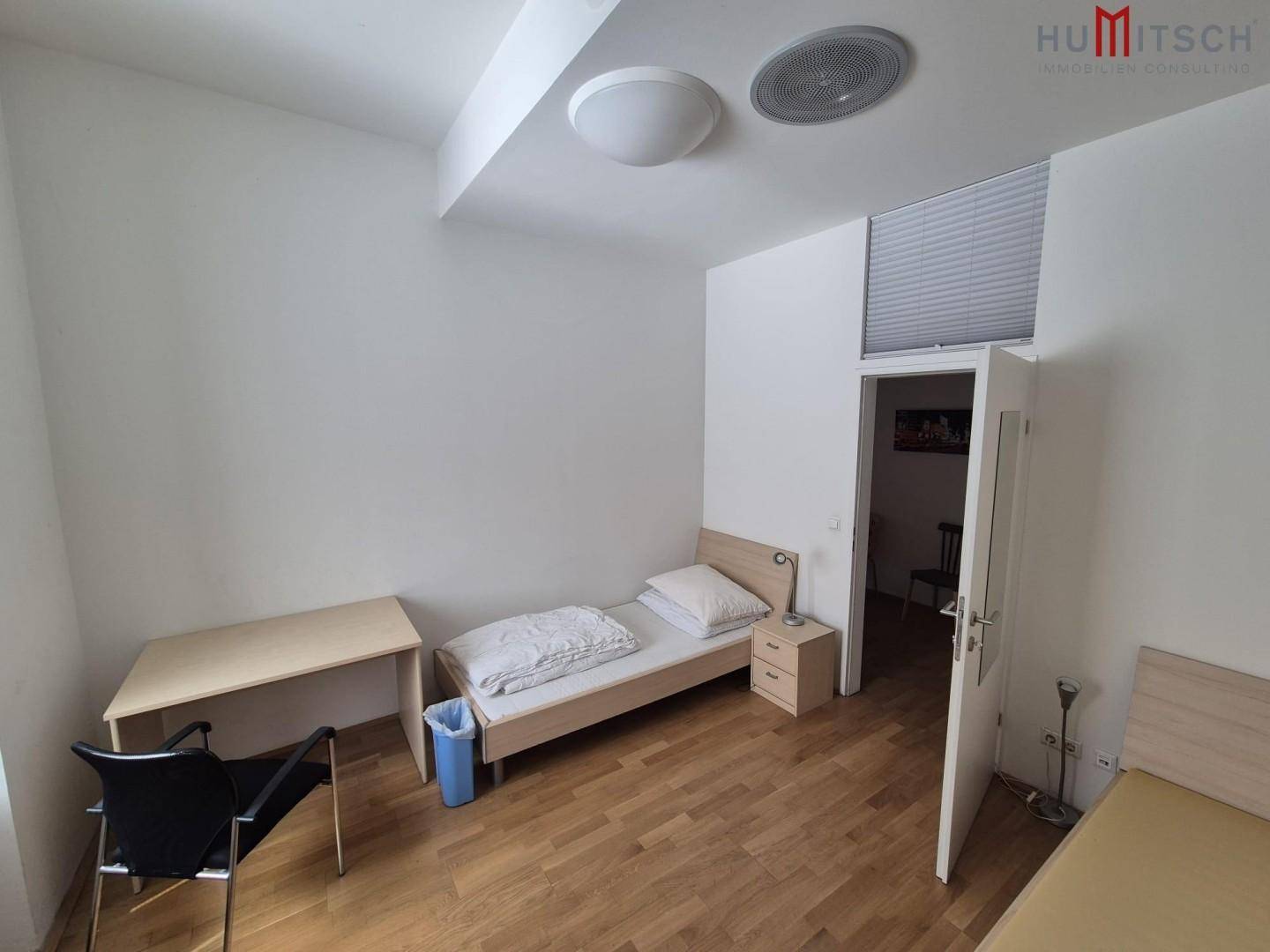 Helles Schlafzimmer mit Einzelbett, Schreibtisch und Nachttisch, mit weiteren Schlafgelegenheiten im Raum.