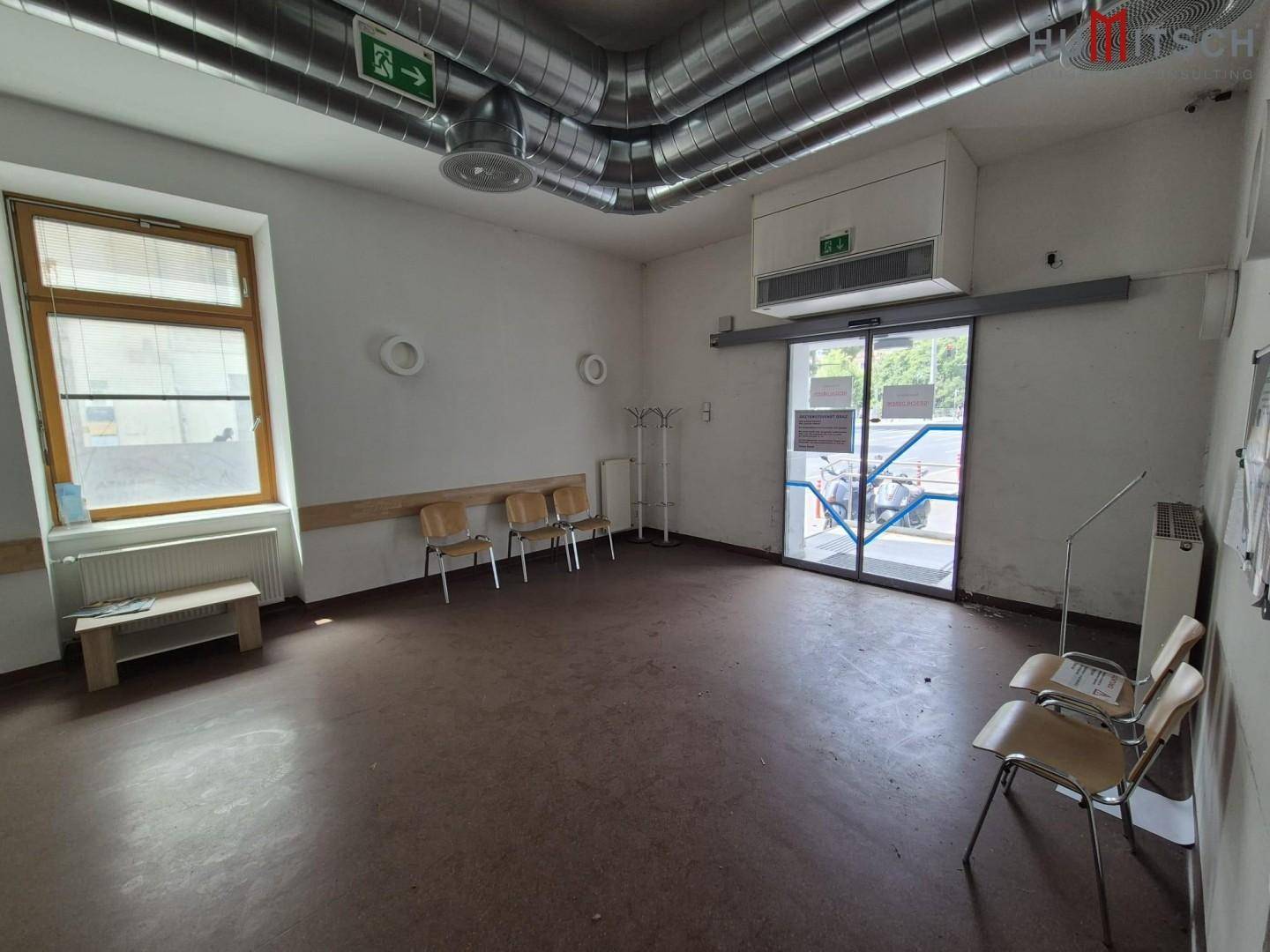 Heller Eingangsbereich und Wartezimmer mit großen Fenstern und sichtbaren Lüftungsrohren.