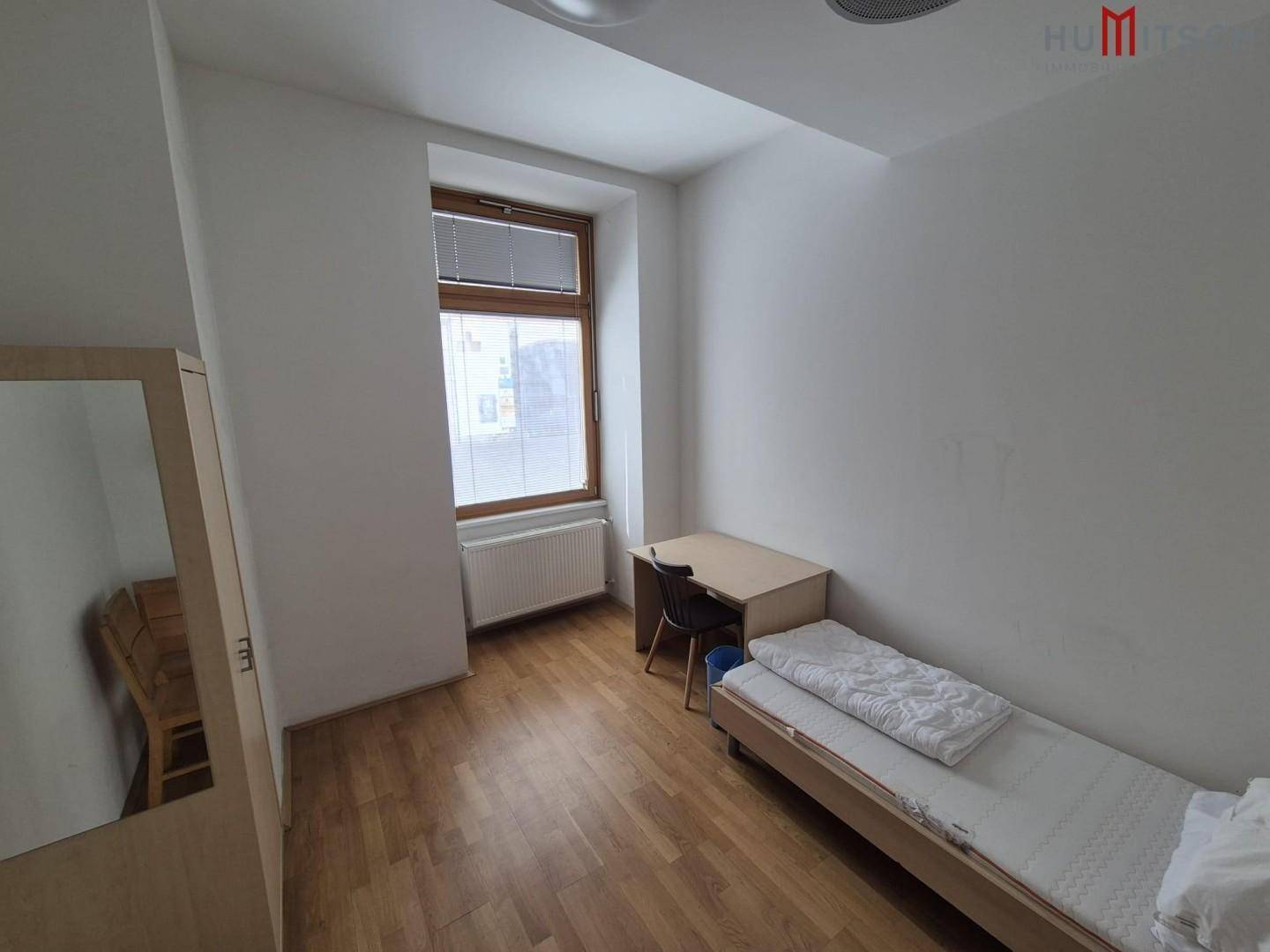 Einfaches Schlafzimmer mit Einzelbett, Schreibtisch und Kleiderschrank, gut beleuchtet durch ein Fenster.