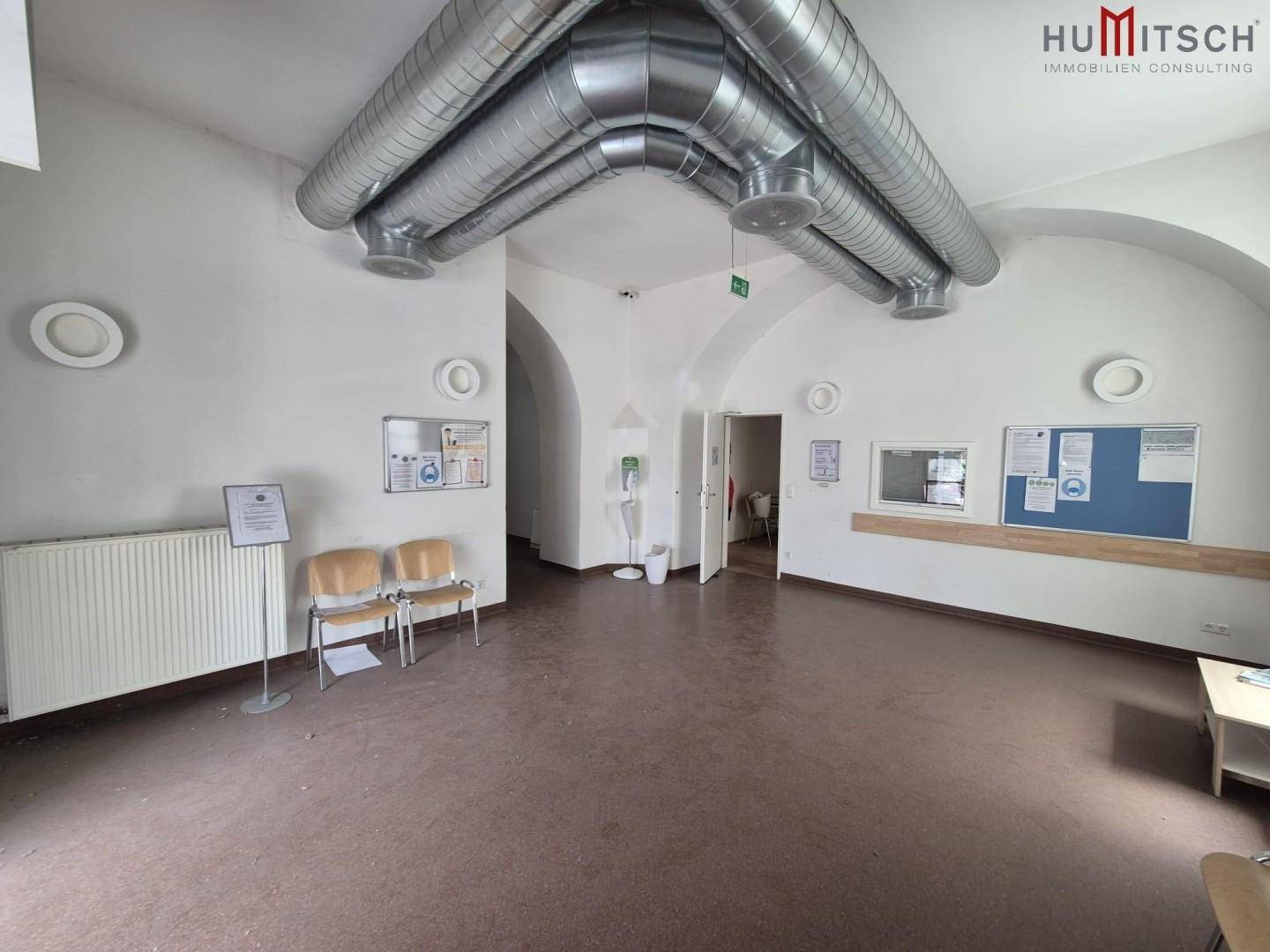 Großer Wartebereich mit sichtbaren Lüftungsrohren und gewölbter Decke, ausgestattet mit Sitzgelegenheiten.