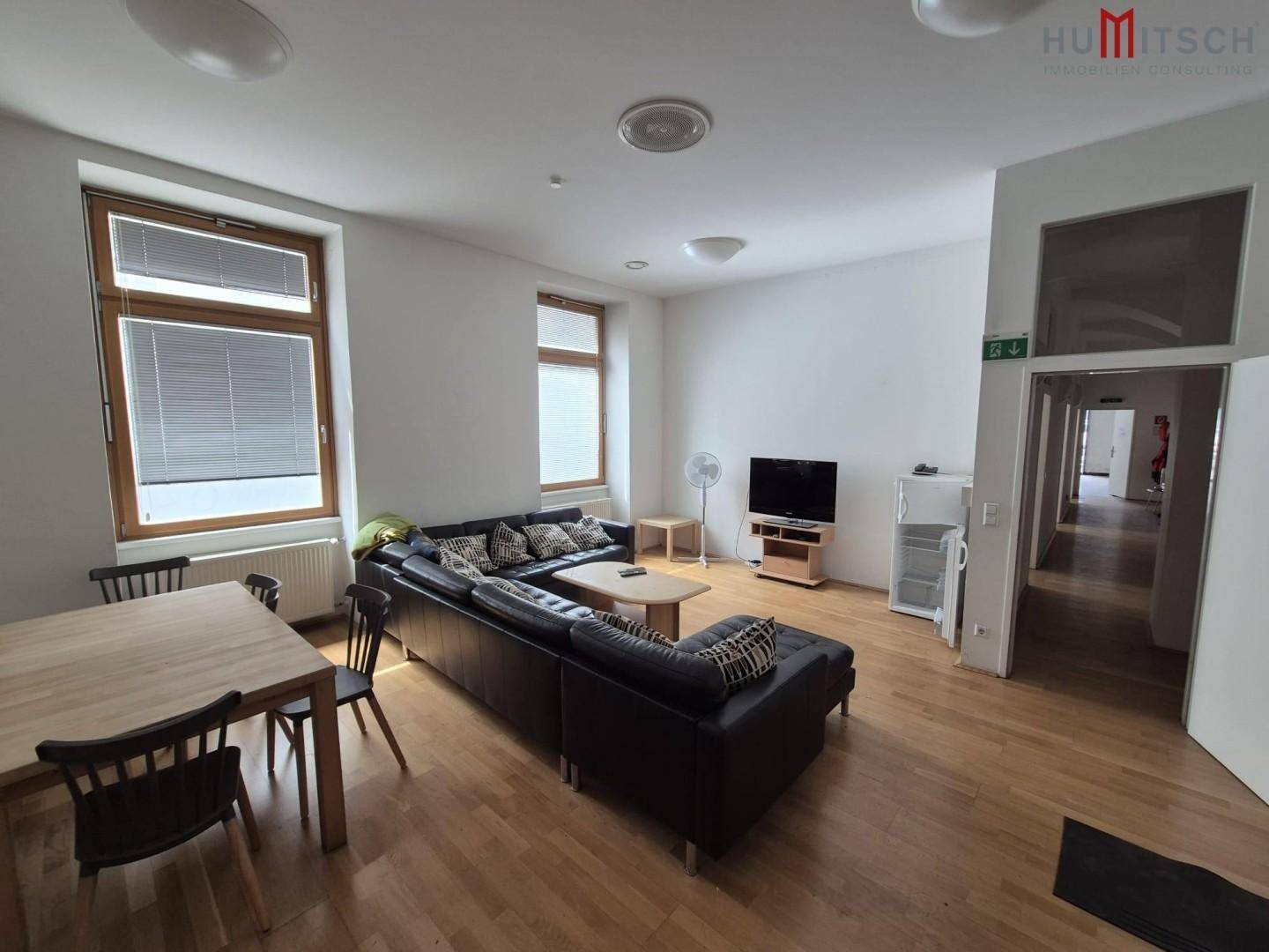 Geräumiger Wohn- und Essbereich mit Holzboden, Sofa und Esstisch, hell durch mehrere Fenster.