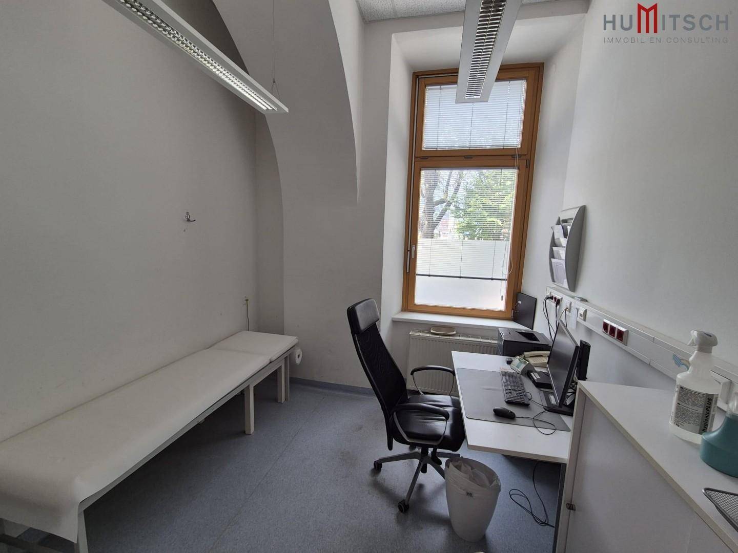 Arztzimmer mit Untersuchungsliege, Schreibtisch und Bürostuhl, gut beleuchtet durch ein großes Fenster.