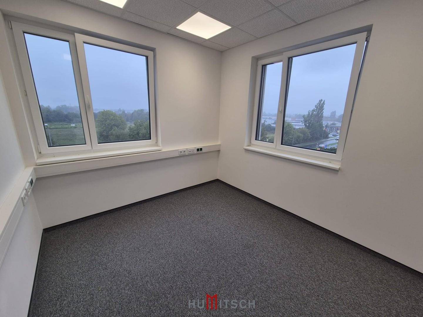 Helles Büro mit Teppichboden und zwei großen Fenstern, die eine weite Aussicht ermöglichen und viel Licht hereinlassen.