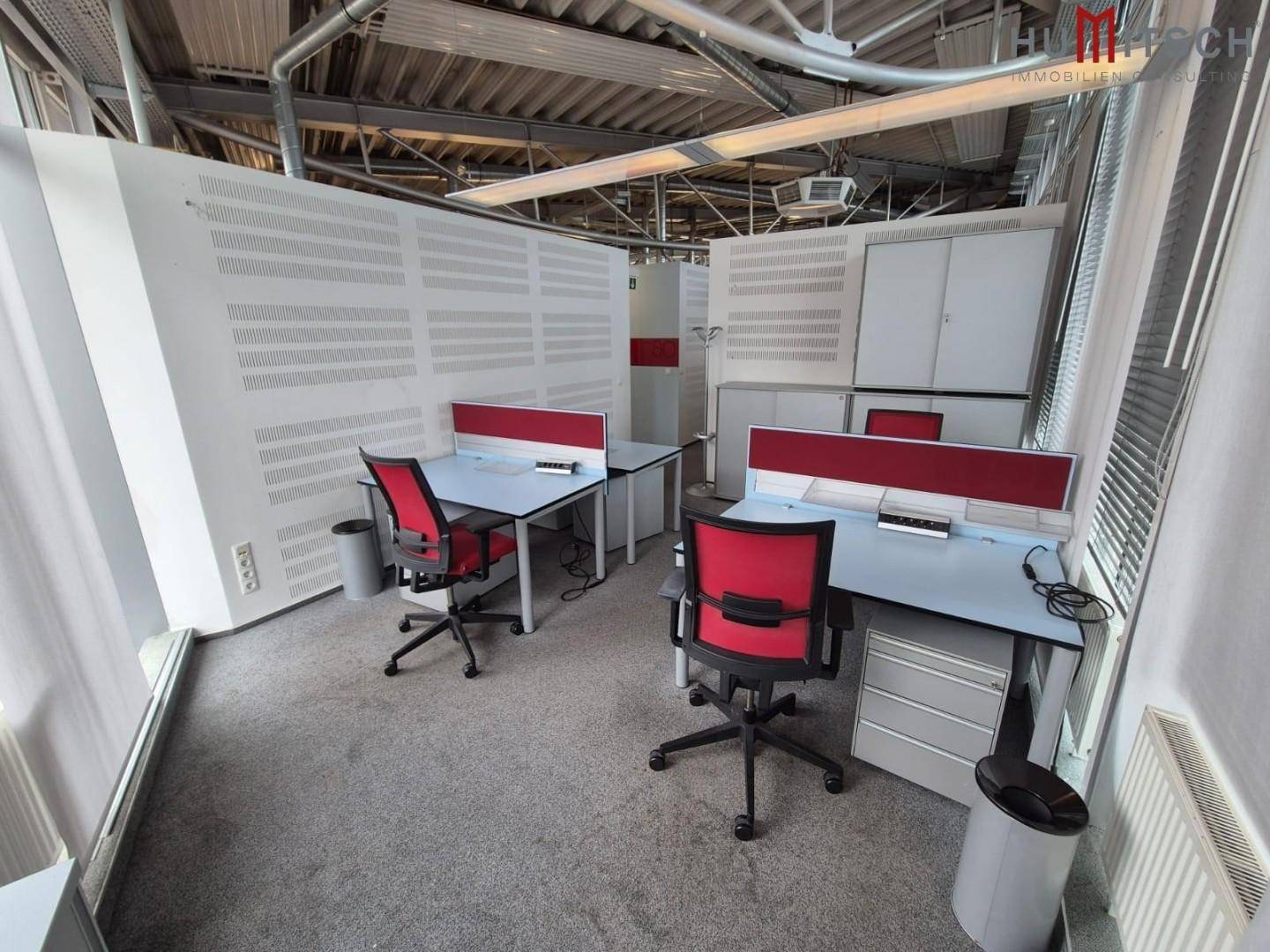 Arbeitsplätze in einem offenen Büro mit Teppichboden, Trennwänden und viel Tageslicht.