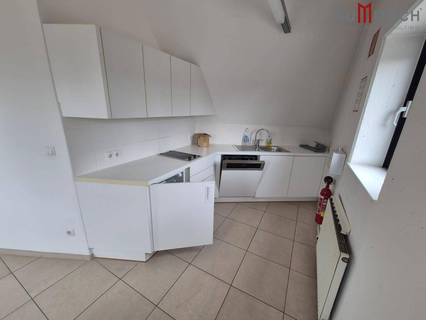 Funktionale Teeküche mit weißen Schränken, Spülmaschine und Fliesenboden unter einer Dachschräge.
