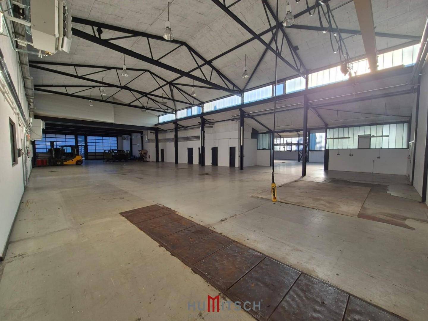 Weitläufige Lagerhalle mit Betonboden, hohen Decken und großen Fenstern, die viel Tageslicht hereinlassen.