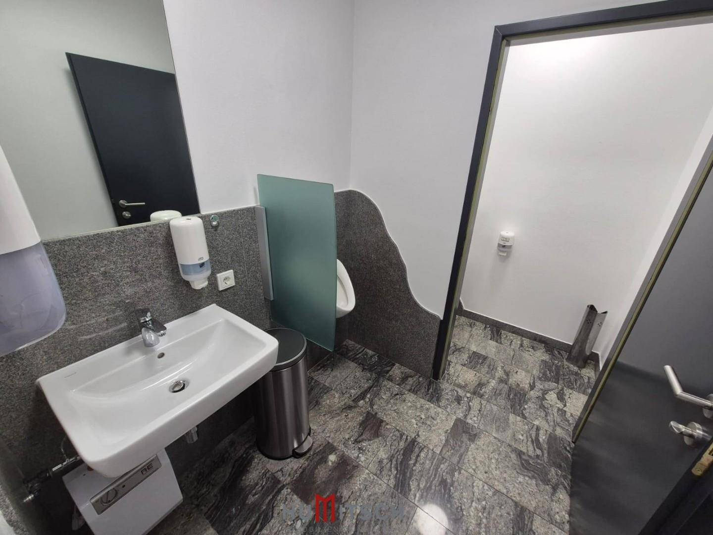 Moderner Herren-WC-Bereich mit Urinal, Waschbecken und grauen Fliesen an Boden und Wand.