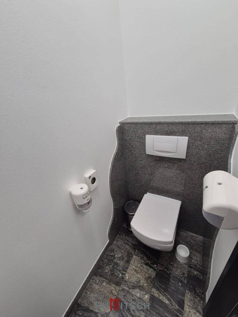 Sauberer Toilettenraum mit wandhängendem WC und dunklen Fliesen, ausgestattet mit Spender.