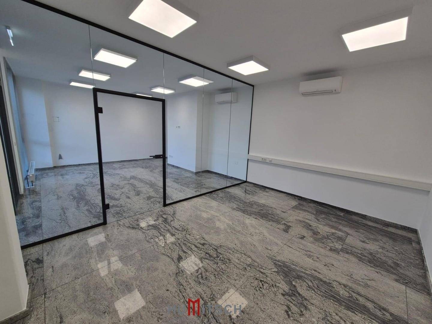 Modernes Büro mit Glaswänden zur Raumtrennung, Fliesenboden und Klimaanlage an der Wand.