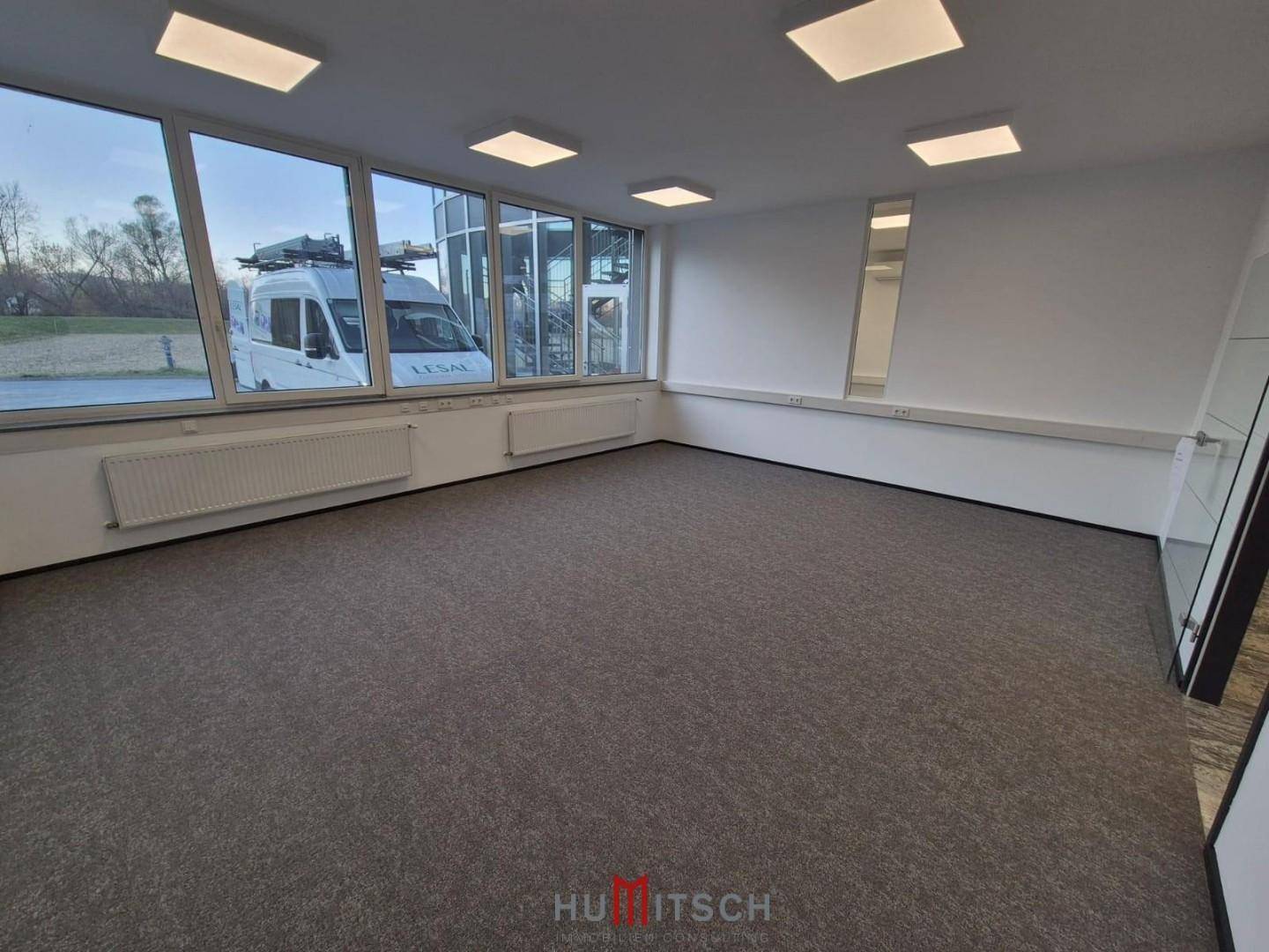 Geräumiges Büro mit großen Fenstern, die viel Tageslicht hereinlassen, und Blick auf den Eingangsbereich.