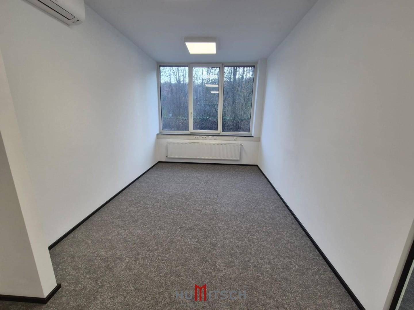 Kleineres Büro mit Teppichboden und einem Fenster, das den Blick ins Grüne freigibt.
