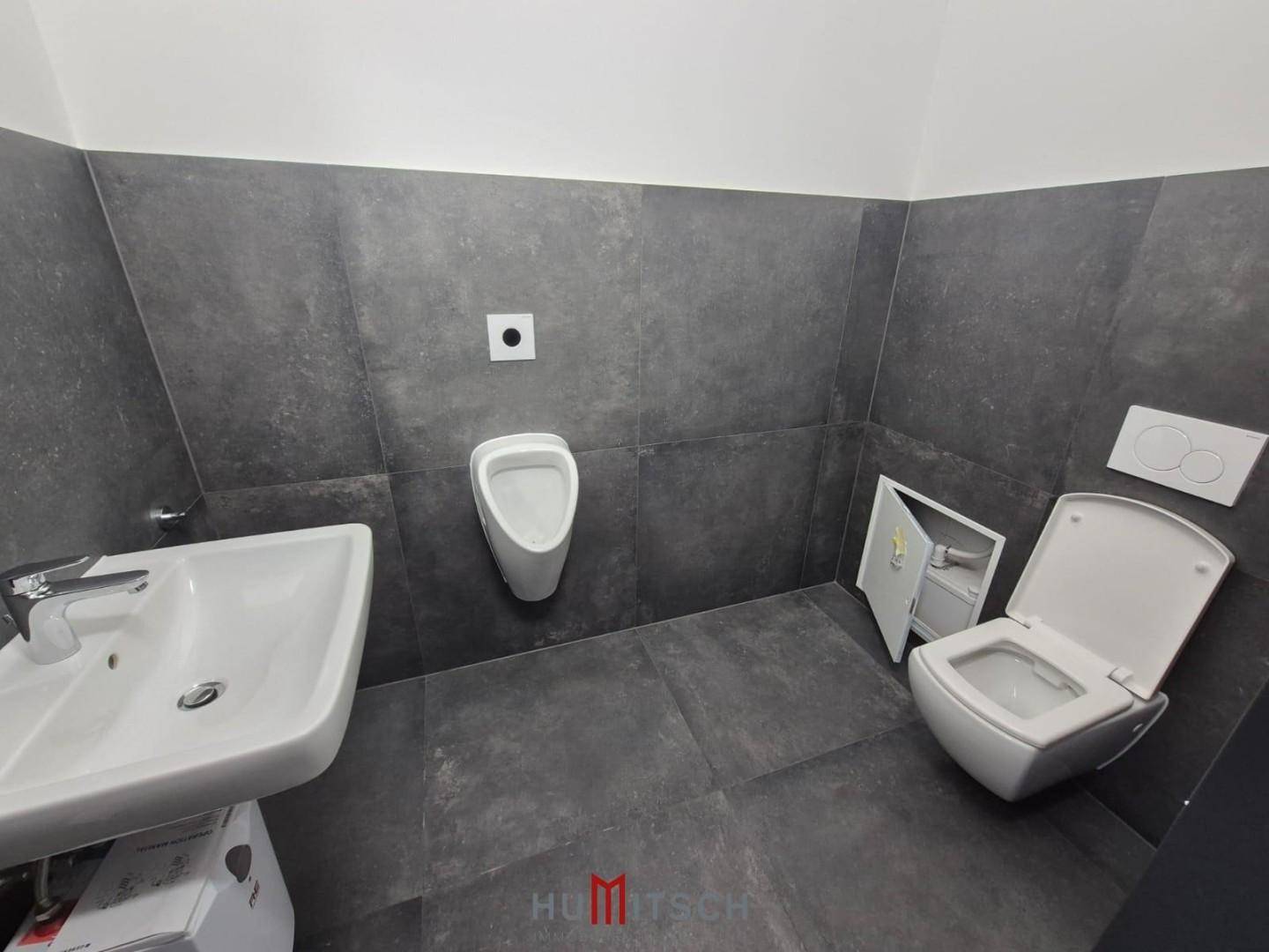 Moderner Toilettenraum mit Waschbecken, Urinal und wandhängendem WC, alles in dunklen Fliesen.