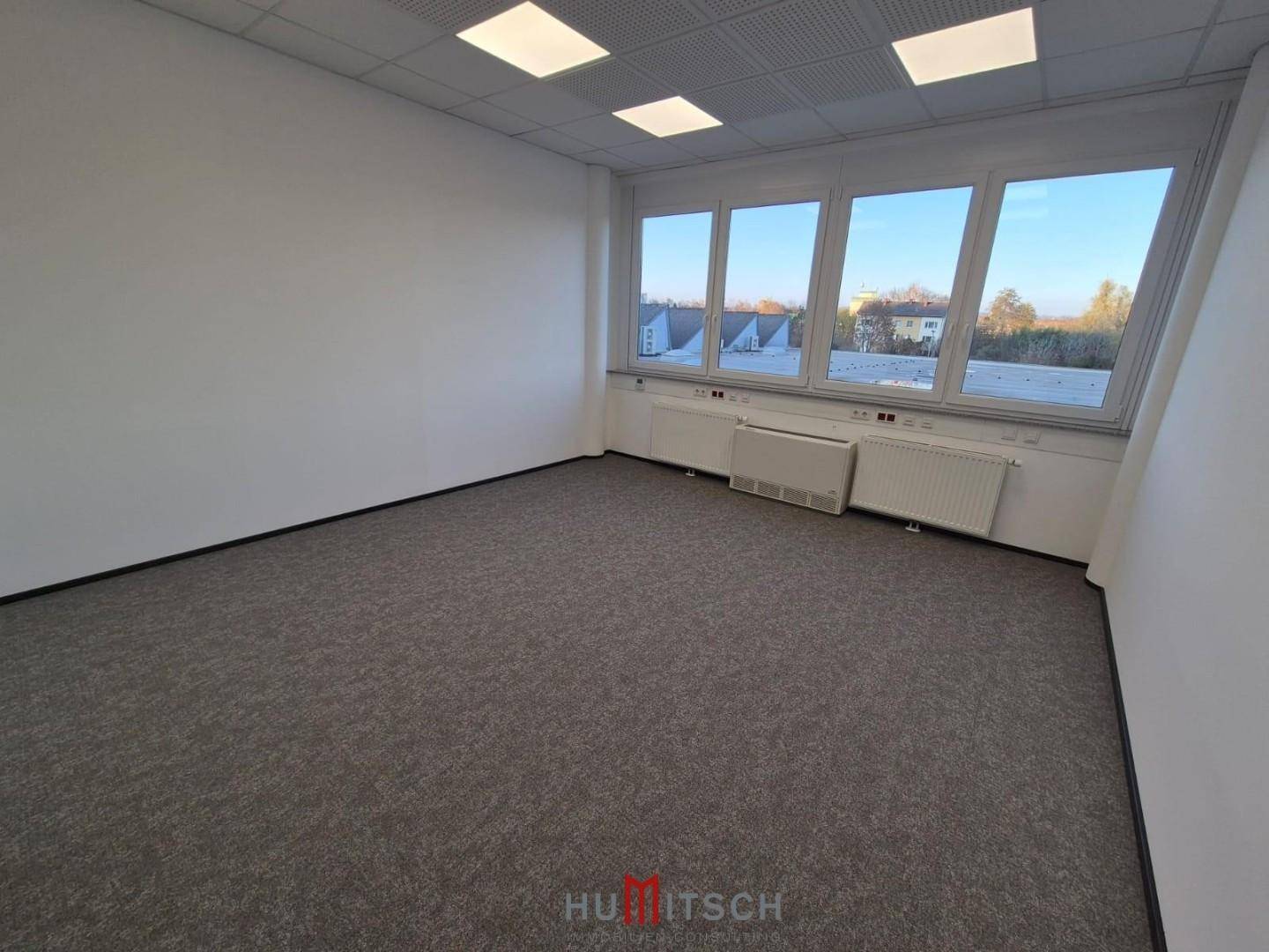 Geräumiges Büro mit Teppichboden und großen Fenstern, die einen weiten Blick auf die Dächer der Stadt bieten.