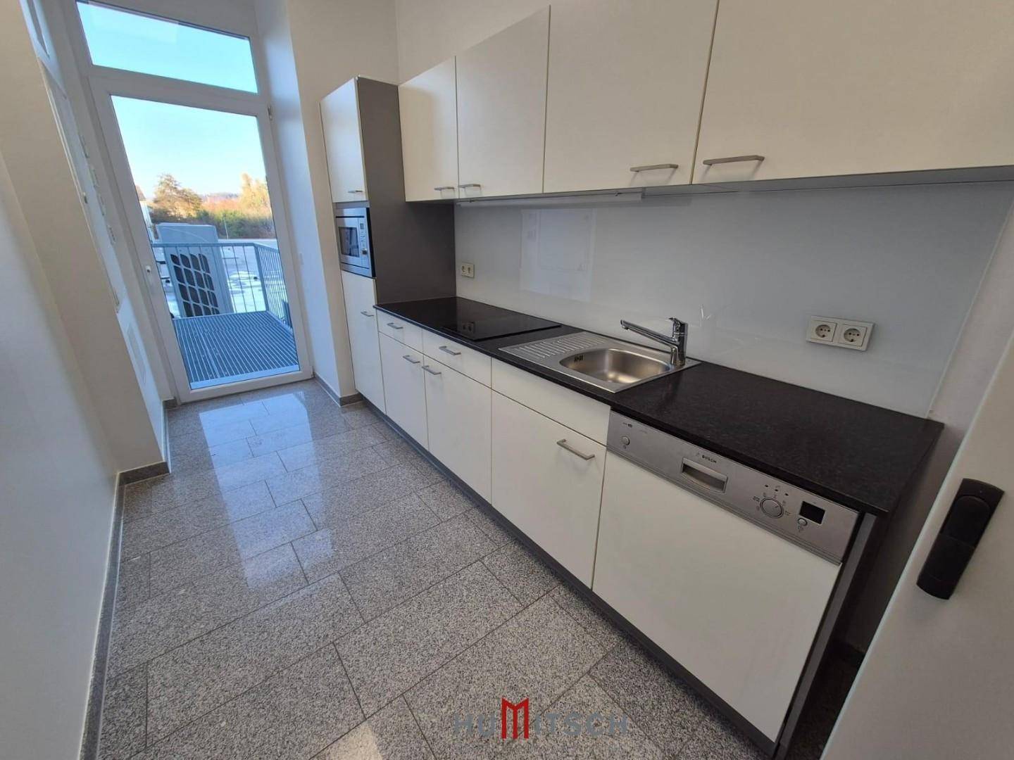 Moderne Küchenzeile mit weißen Schränken, dunkler Arbeitsplatte und Zugang zu einem kleinen Balkon.