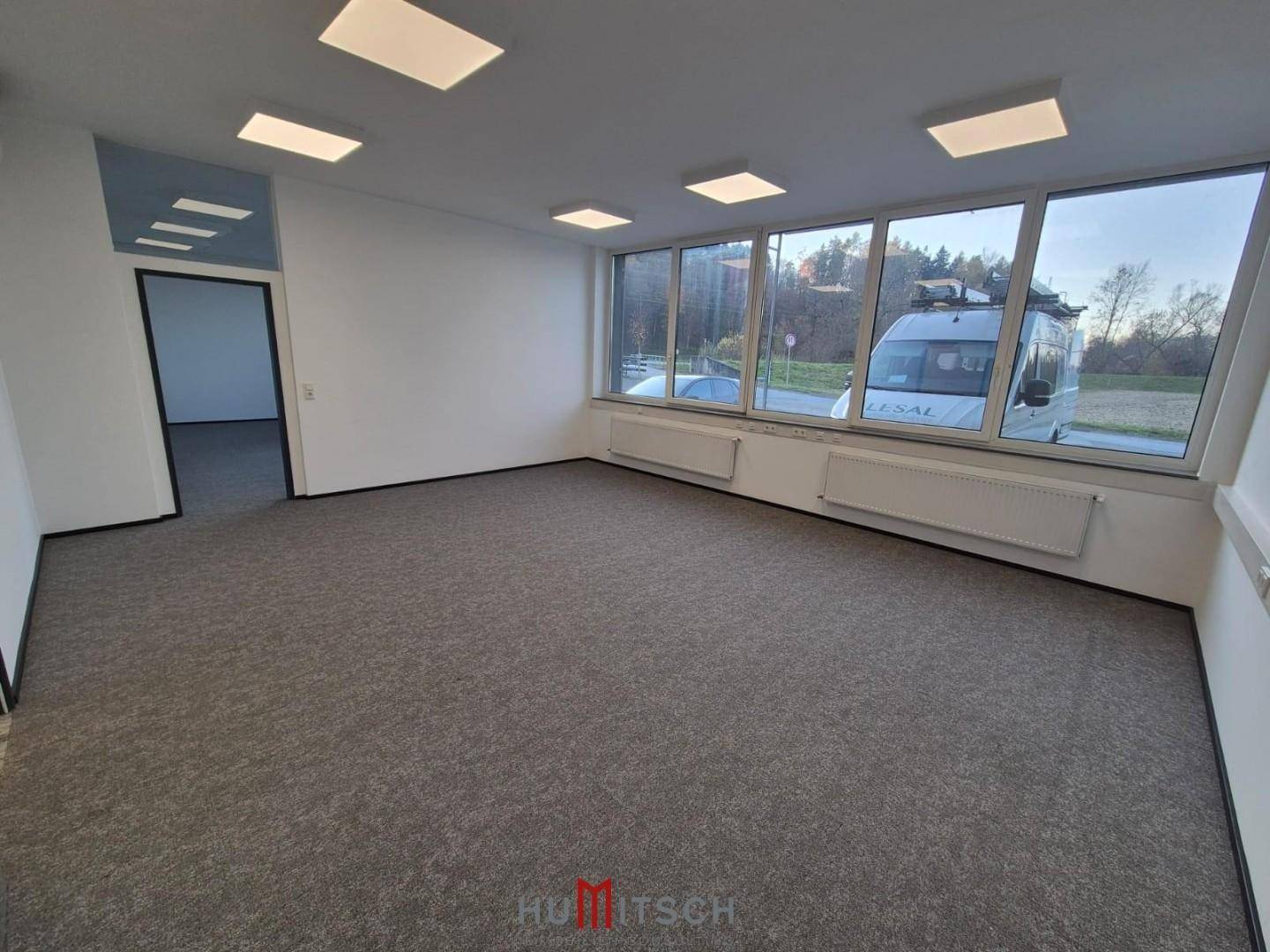 Helles Büro mit Teppichboden, großen Fenstern und Blick auf die Straße und umliegende Landschaft.