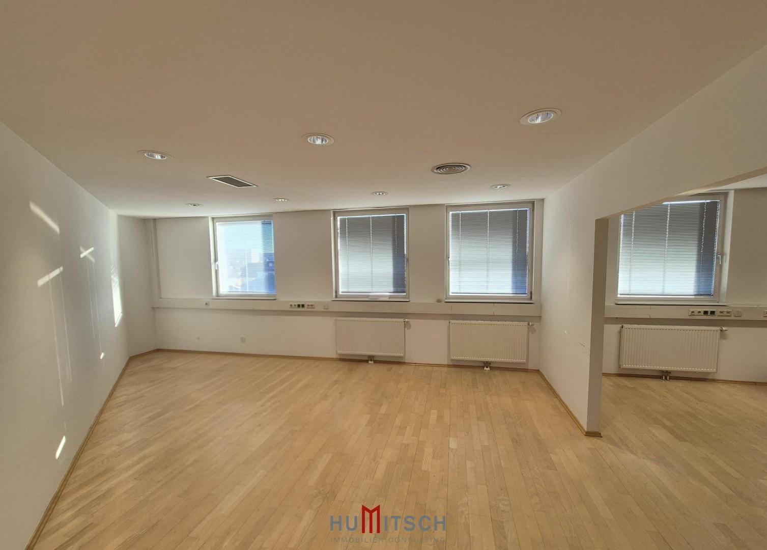 Leeres Büro mit hellem Parkettboden, Heizkörpern unter den Fenstern und Jalousien für Privatsphäre und Sonnenschutz.