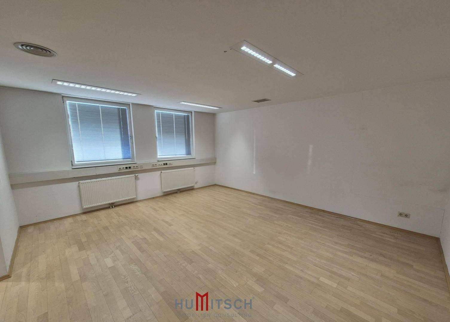 Geräumiges, leeres Büro mit Holzparkett, großen Fenstern und integrierter Deckenbeleuchtung, bereit zur Einrichtung.