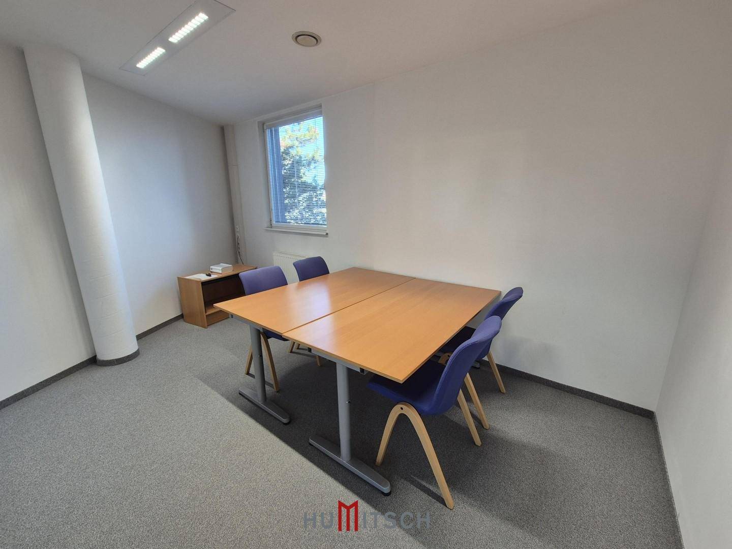 Kleiner Besprechungsraum oder Einzelbüro mit Holztisch und blauen Stühlen, gut beleuchtet.