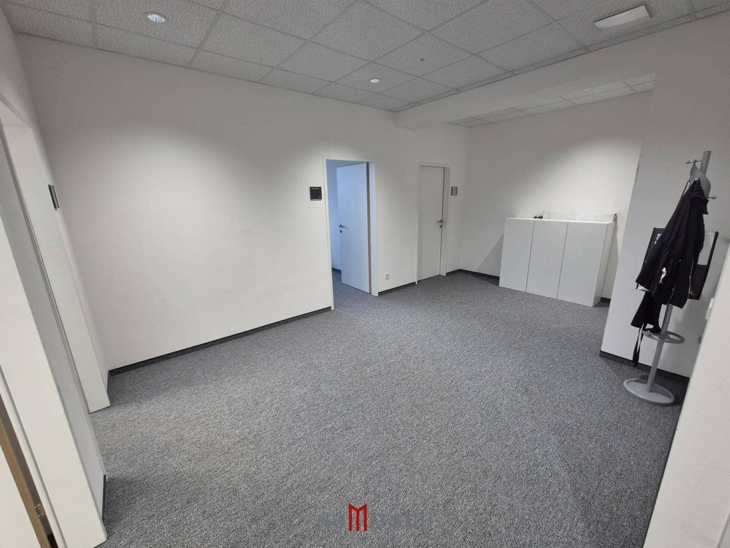Leerer Büroraum mit Teppichboden, weißen Wänden und einer Garderobe am Eingangsbereich.