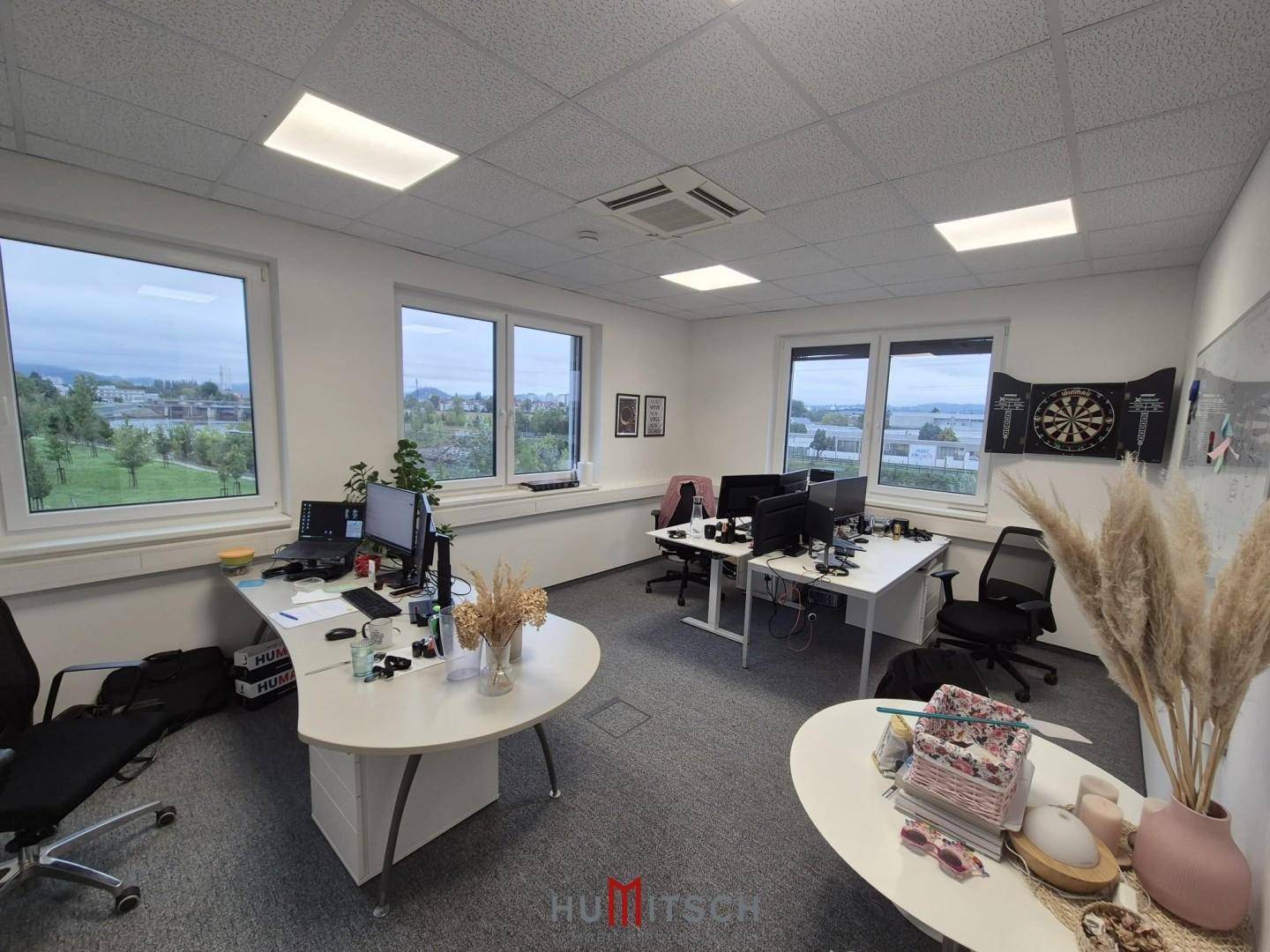 Offenes Büro mit mehreren Schreibtischen, Teppichboden und großen Fenstern, die viel Licht hereinlassen.