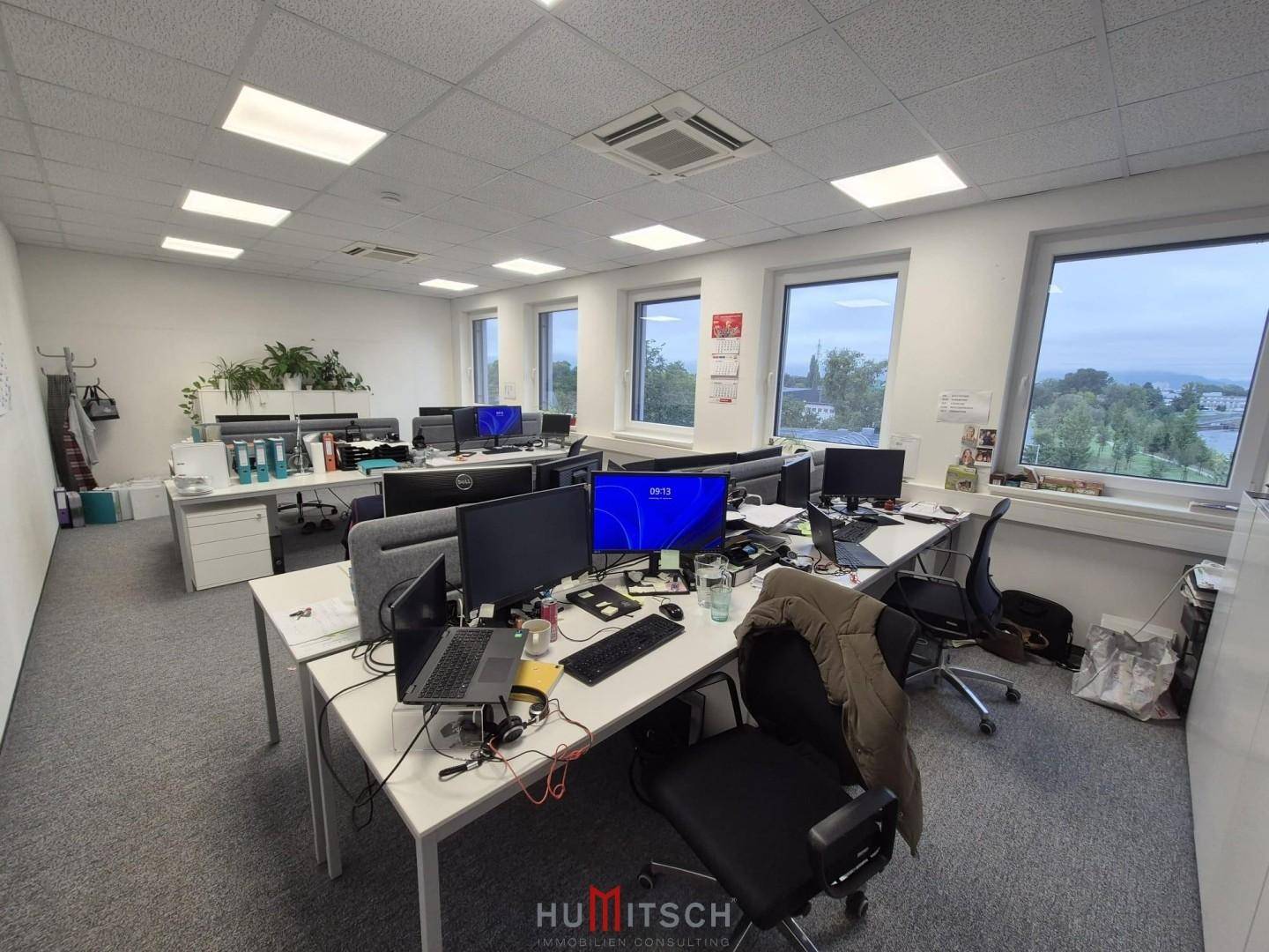 Großes Büro mit vielen Arbeitsplätzen, Monitoren und Blick auf die grüne Umgebung.