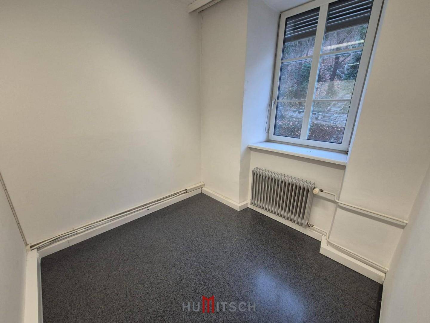 Kleinerer, heller Raum mit grauem Boden, weiß gestrichenen Wänden und einem Fenster mit Heizkörper.