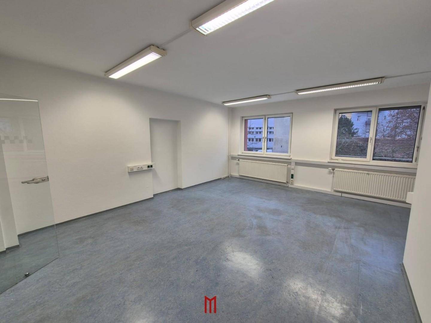 Geräumiges, leeres Büro mit großen Fenstern, grauem Boden und modernen Deckenleuchten.