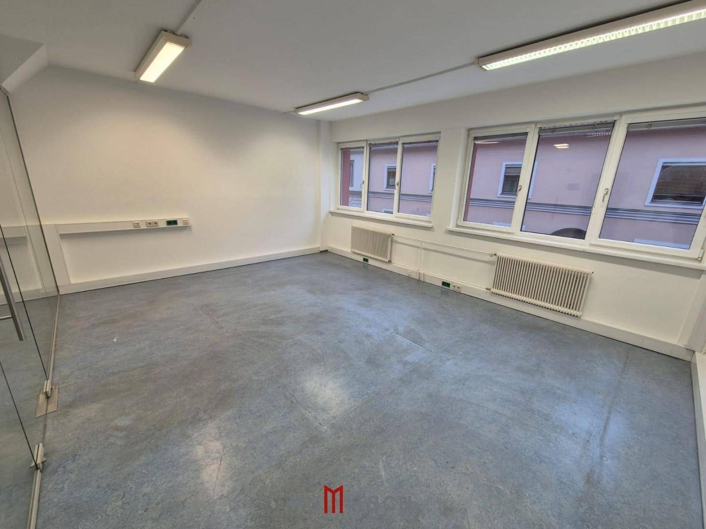 Modernes Büro mit Glaswand, großen Fenstern und grauem Bodenbelag, ideal für Arbeitsbereiche.