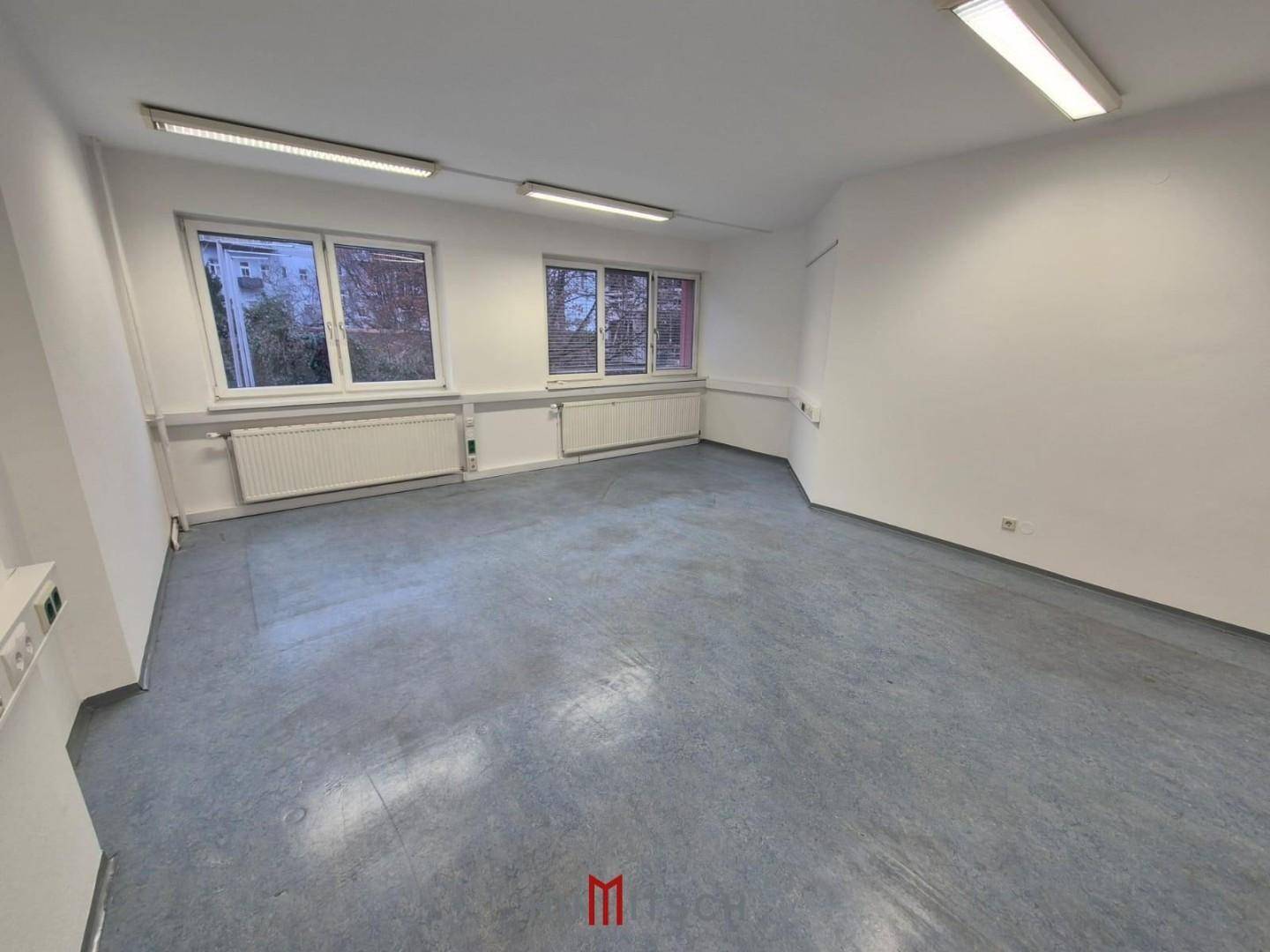 Helles Eckbüro mit zwei Fenstern, weiß gestrichenen Wänden und grauem Bodenbelag.